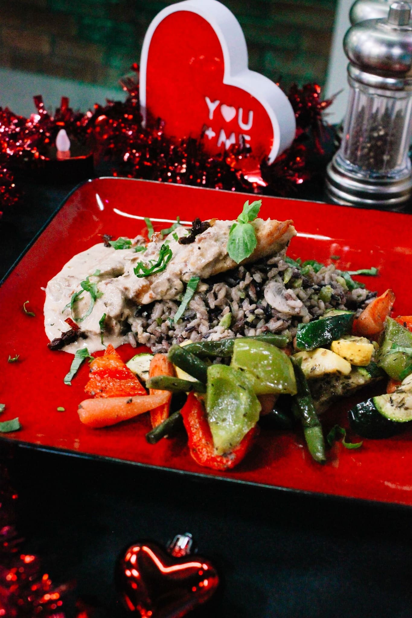 A festive meal on a red plate features chicken in creamy sauce over rice with sautéed carrots, zucchini, and peppers. Valentine’s Day decor, red tinsel, and a heart with YOU + ME are in the background.