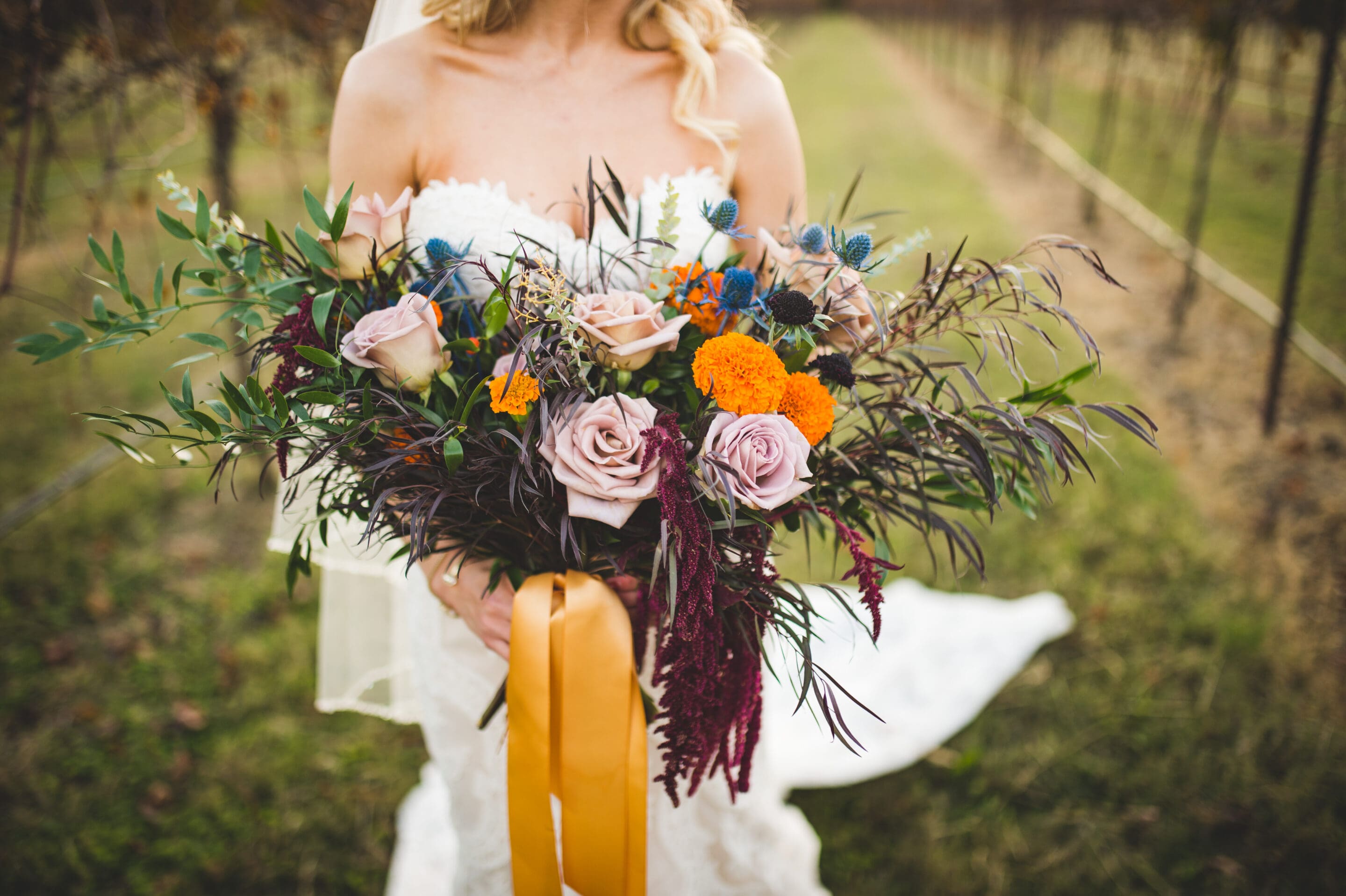 A bride in a white dress holds a large bouquet of colorful flowers, including roses, marigolds, and greenery, tied with a yellow ribbon, standing outdoors in a grassy, vineyard-like setting.
