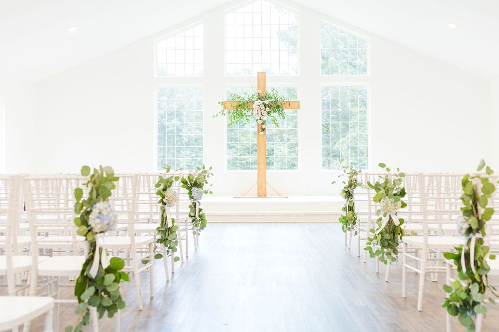 Bright, airy chapel with white chairs lined up on each side of the aisle, decorated with greenery. At the front, a large wooden cross adorned with flowers, stands before tall windows letting in natural light.