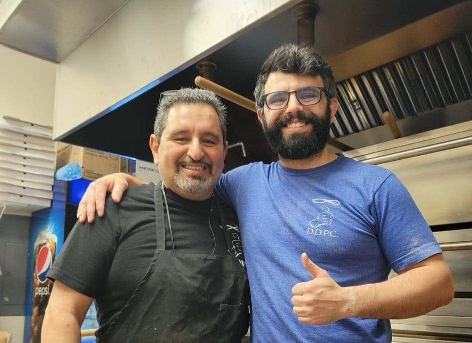 Two men stand together in a kitchen, smiling at the camera. One wears a black apron and the other, in a blue shirt, gives a thumbs up. Stainless steel kitchen equipment and a Pepsi machine are visible in the background.