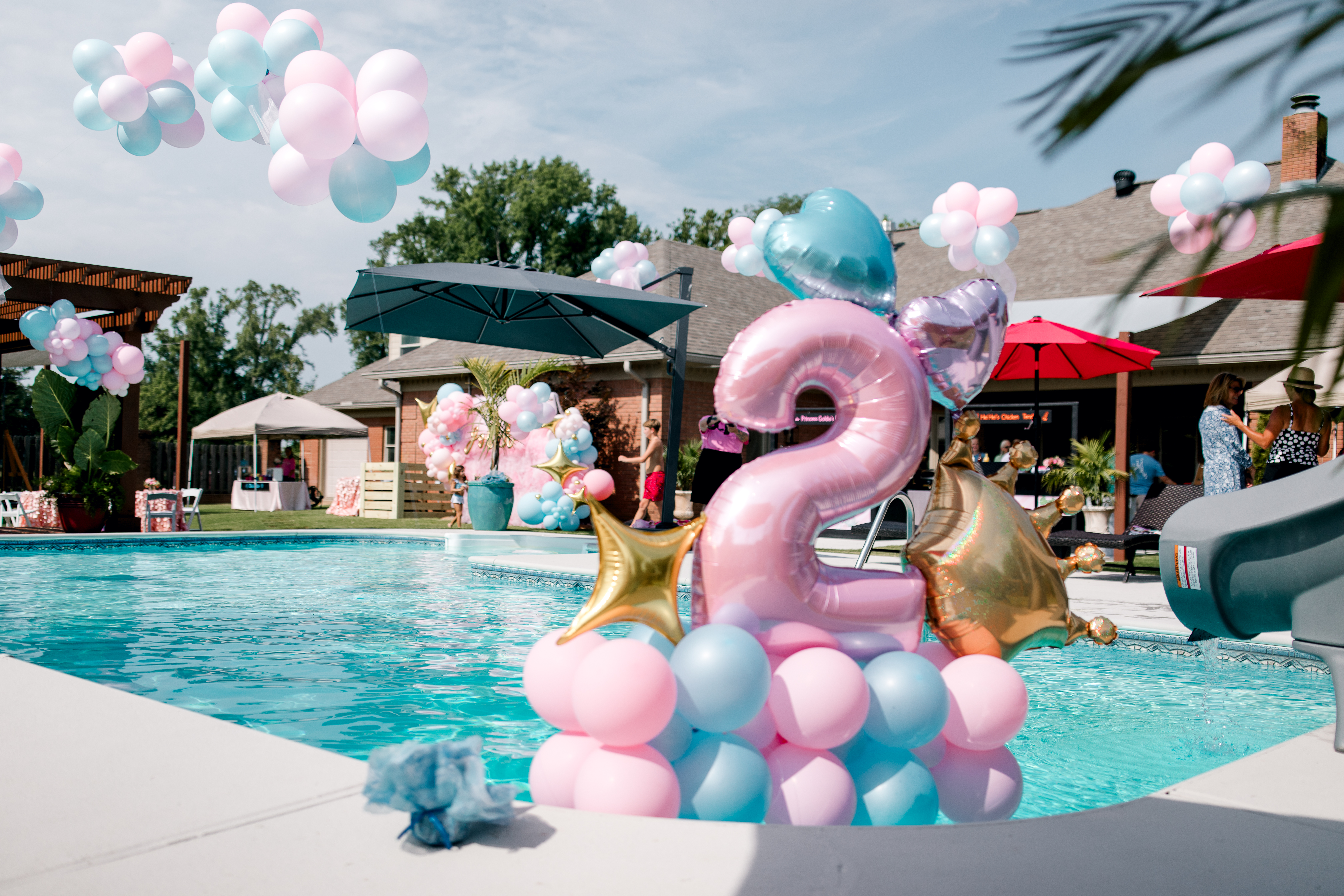 A backyard pool decorated with colorful balloons, a large pink number 2, and gold stars sets the scene for a Poolside Princess Party. Festive food and party fun await, with people, umbrellas, and a slide under the blue sky.