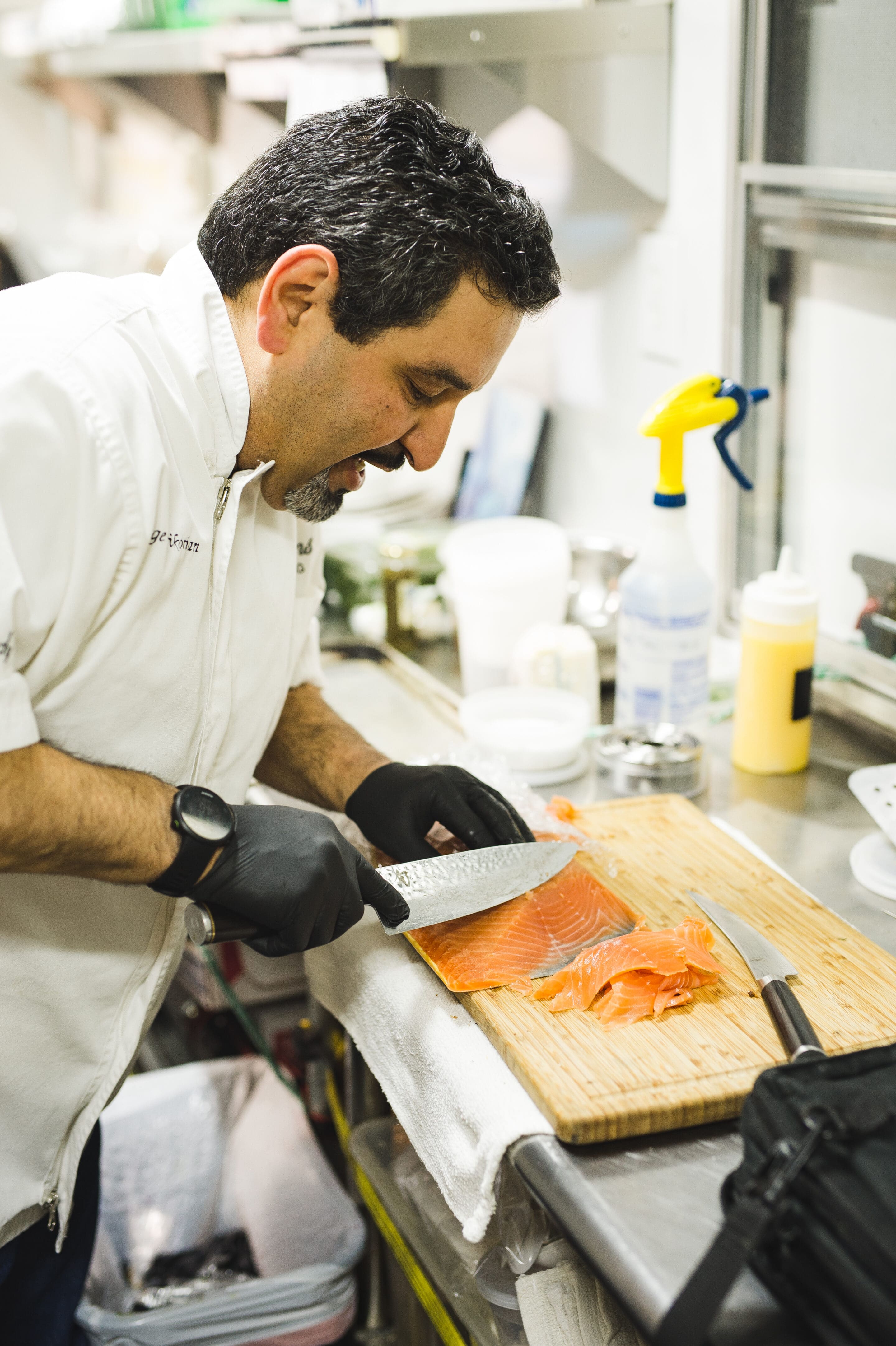 A chef wearing a white coat and black gloves slices a fillet of salmon on a wooden cutting board in a commercial kitchen. Various kitchen tools and containers are visible in the background.