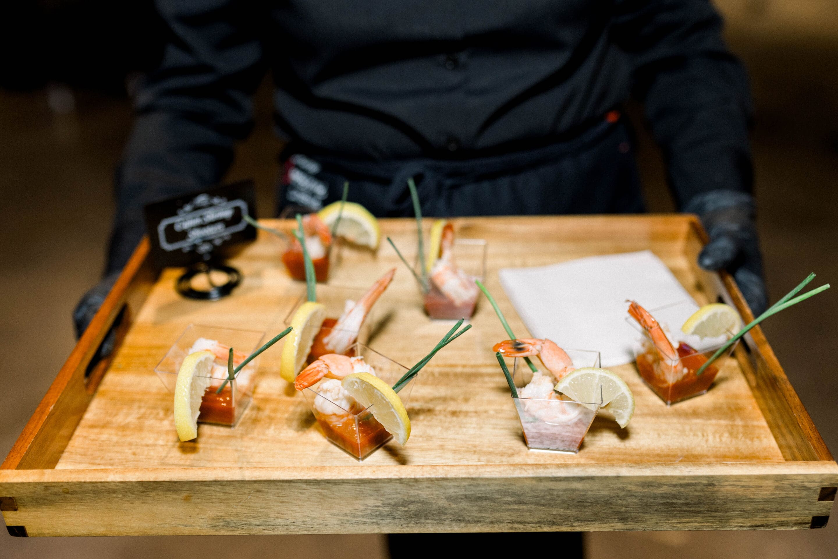 A server in black attire holds a wooden tray with cocktail shrimp in small clear cups, garnished with lemon slices and green chives, alongside cocktail sauce and neatly folded napkins.