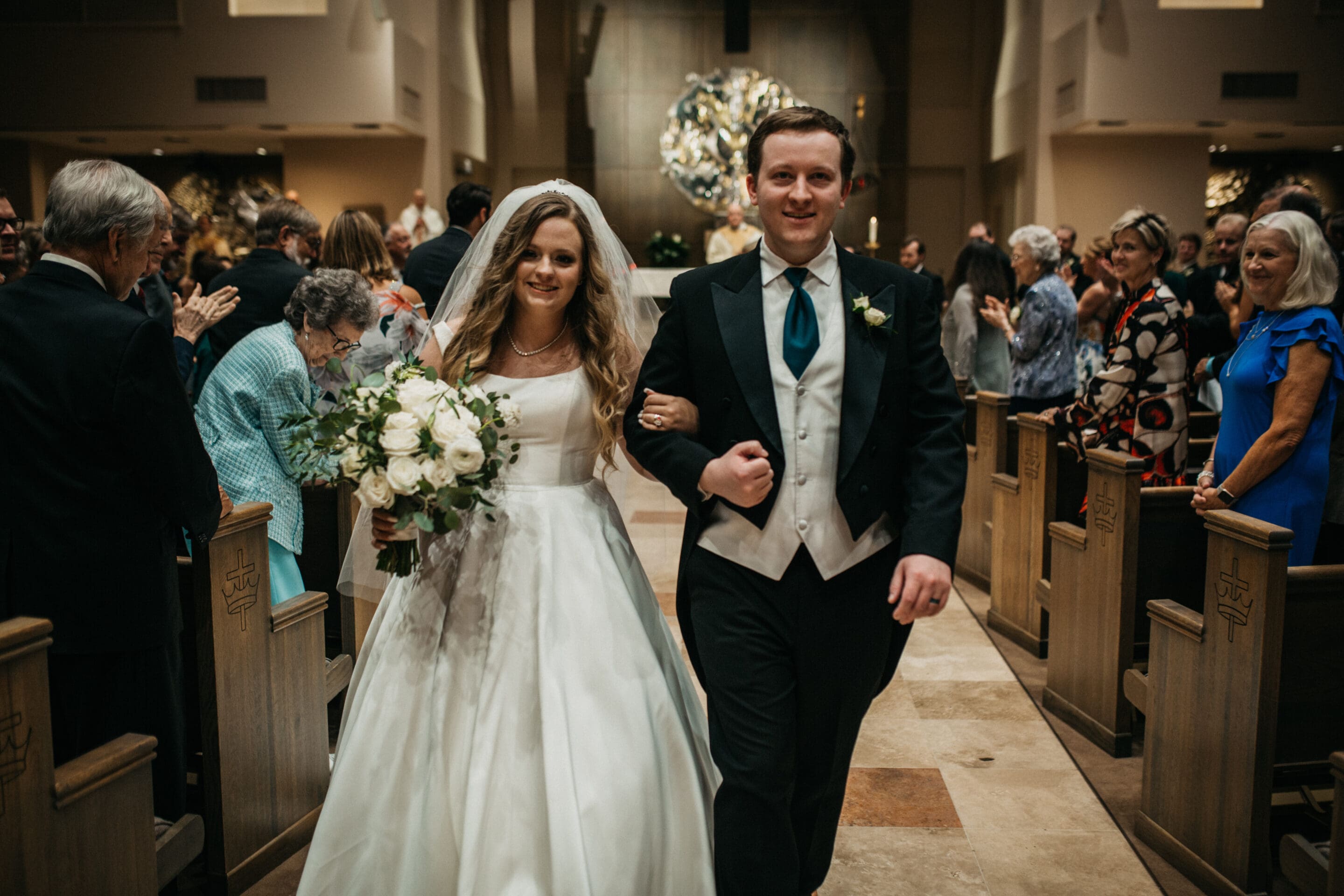 A bride in a white dress and a groom in a tuxedo walk arm in arm down the aisle of a church, smiling, with guests standing and watching on either side.