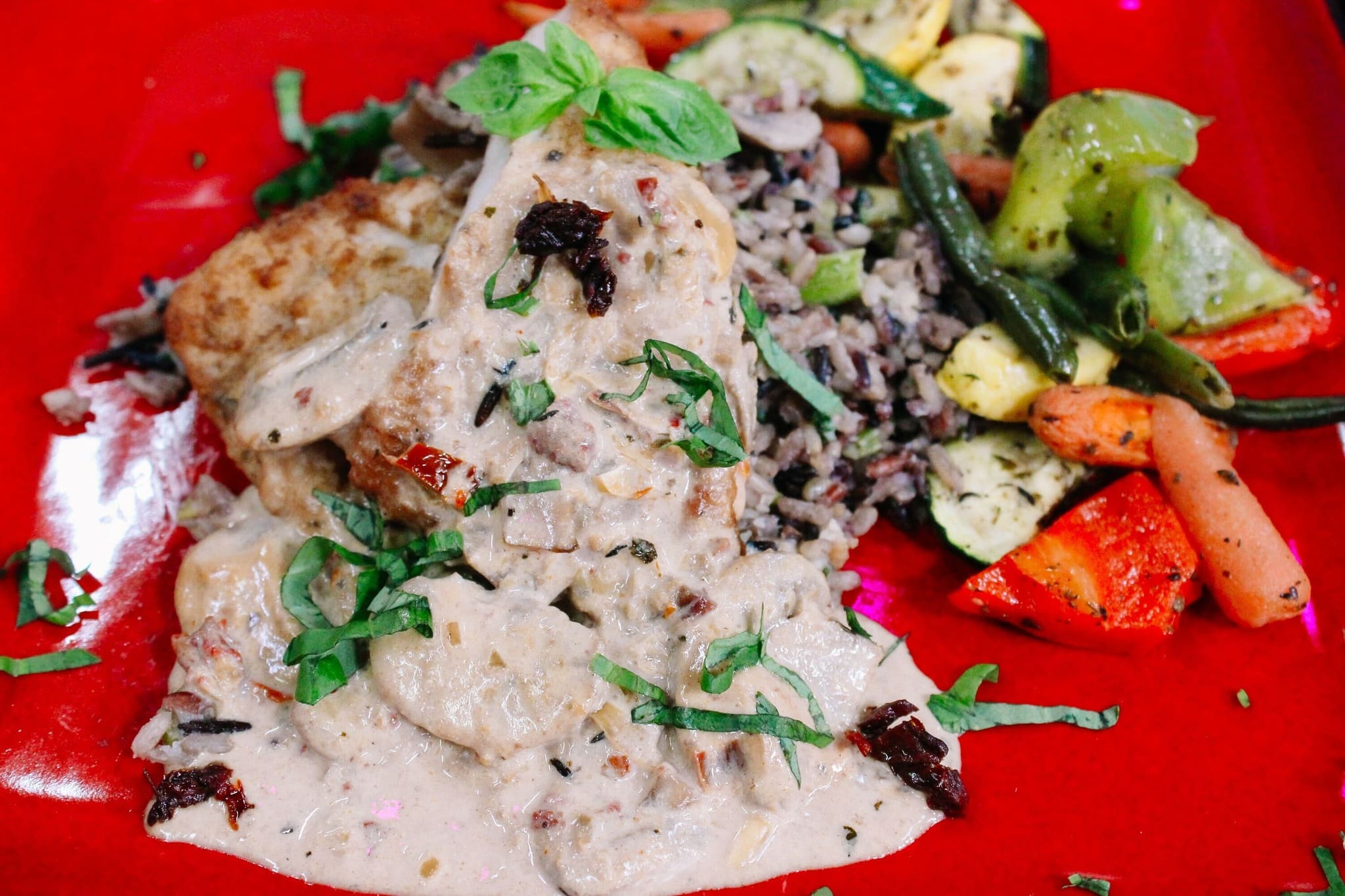 A plate of creamy chicken with herbs, served over wild rice with sautéed mixed vegetables including carrots, zucchini, and green beans, garnished with fresh basil on a red plate.