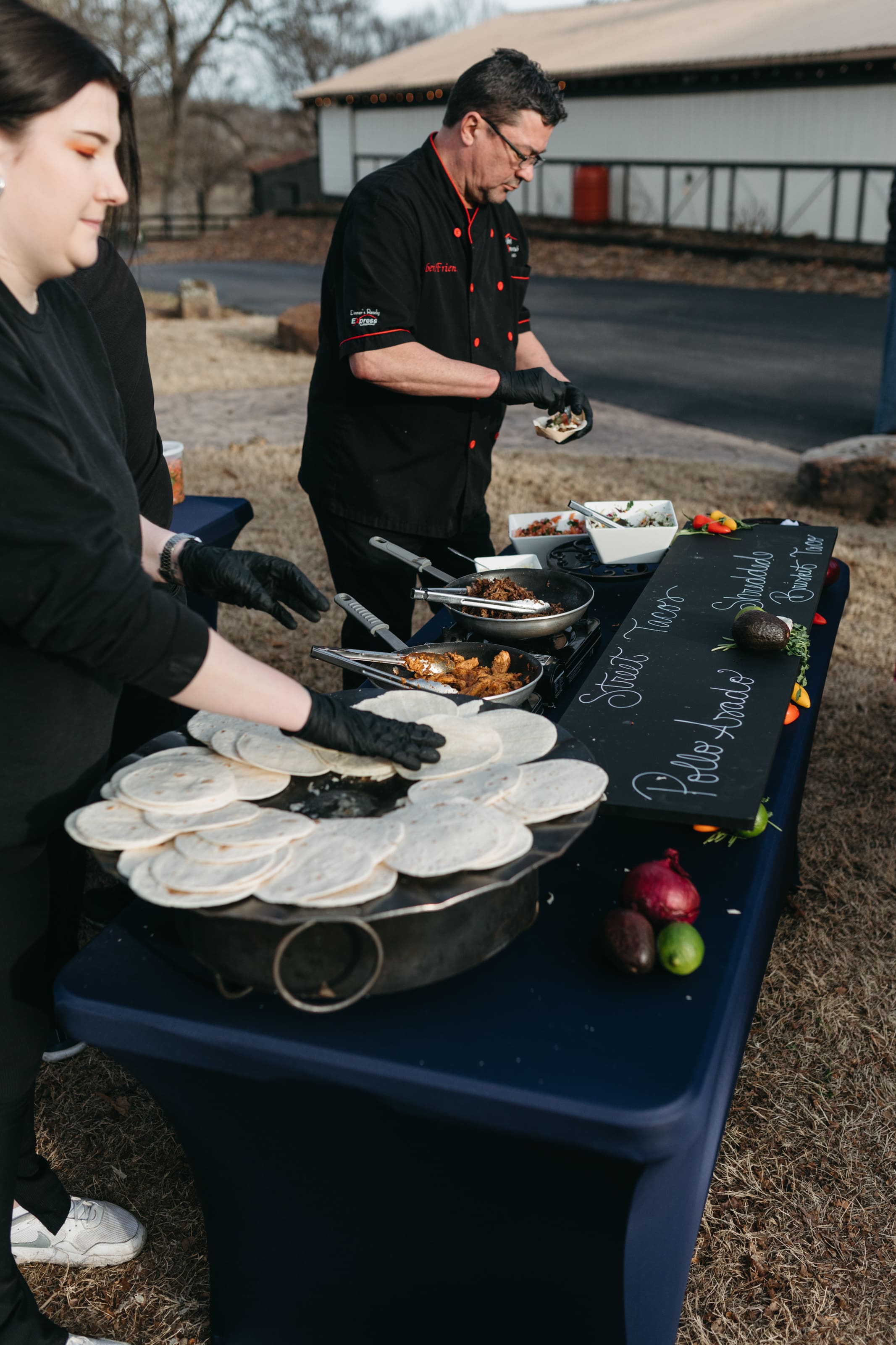 Two people prepare tacos at an outdoor food station with stacks of tortillas, various fillings, and a chalkboard menu. Both wear black clothing and gloves, and the scene is set on a grassy area near a building.