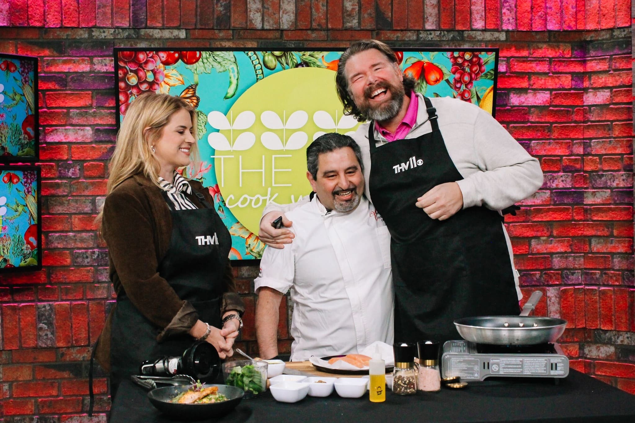 Three people stand smiling in a colorful kitchen studio. The man in the middle wears a chefs coat, while the others wear aprons. Cooking ingredients and utensils are on the counter in front of them.