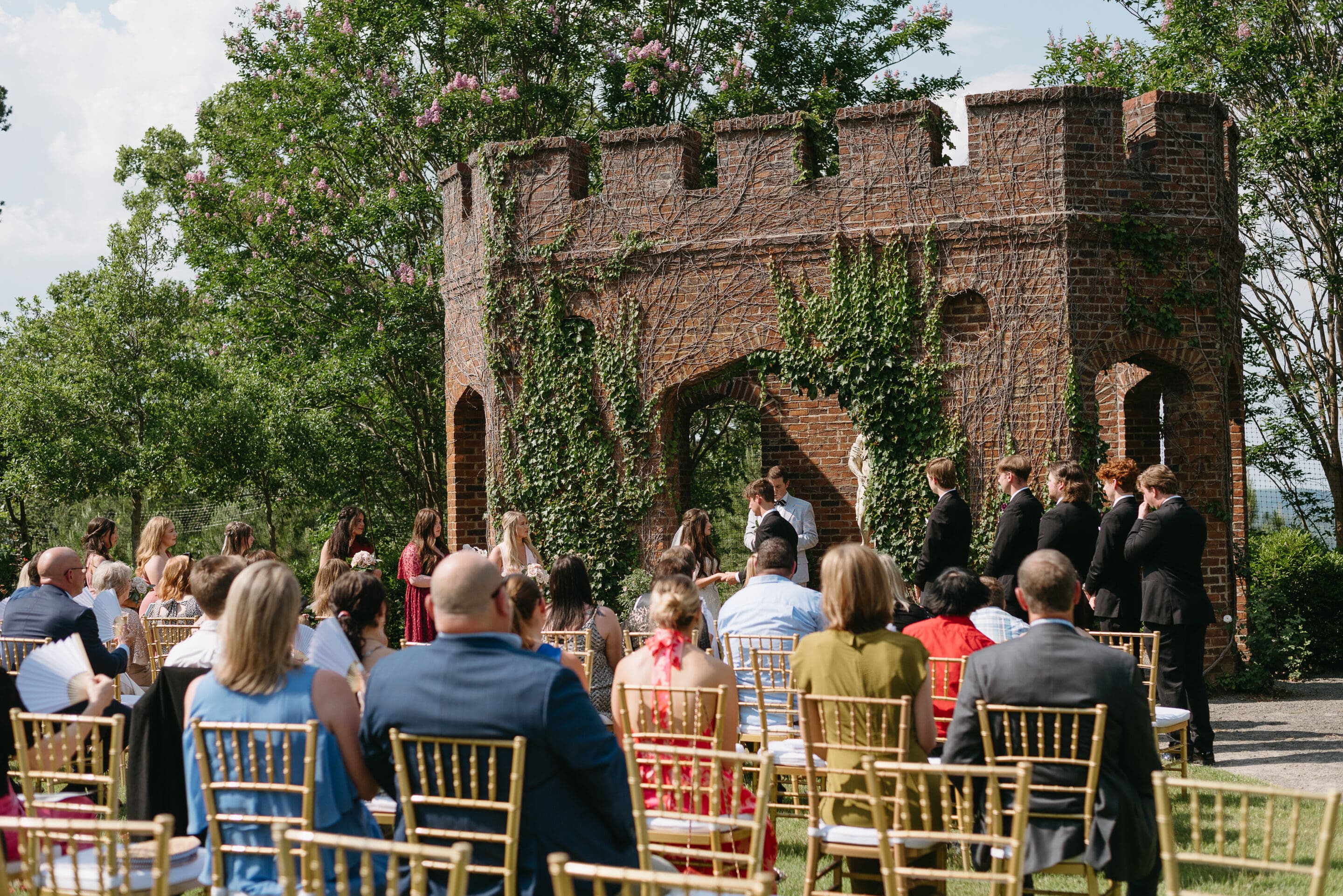 An outdoor wedding ceremony with guests seated on gold chairs facing a couple and officiant in front of a brick, ivy-covered castle-like structure, surrounded by greenery on a sunny day.
