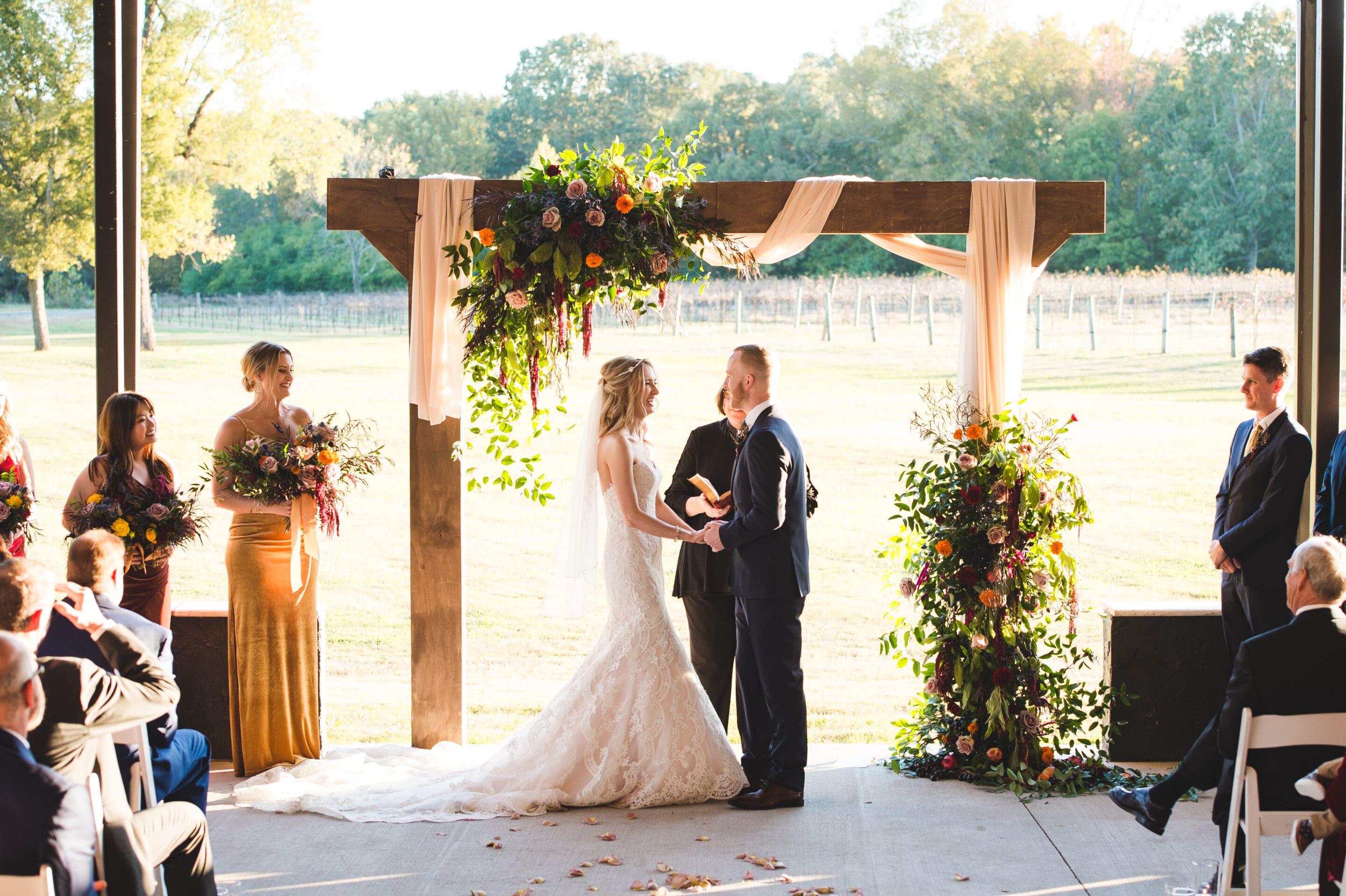 A bride and groom stand holding hands under a wooden floral arch during an outdoor wedding ceremony. Bridesmaids and groomsmen stand nearby, and guests are seated, facing the couple. Sunlight streams in from the field behind them.