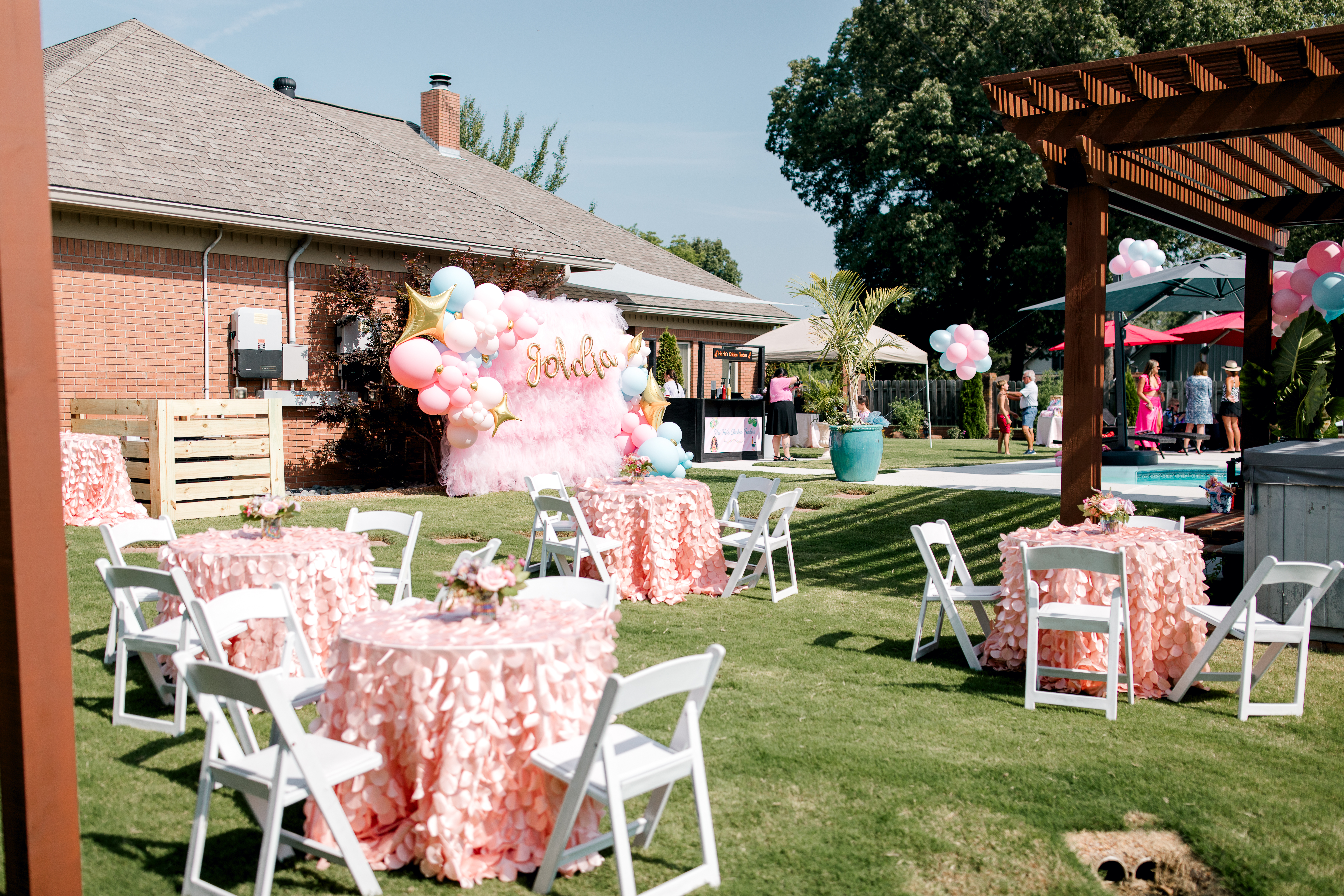 The image captures a backyard celebration, possibly a party, with several round tables draped in light pink ruffled tablecloths and surrounded by white folding chairs on a green lawn. A pink and white balloon arch with gold lettering stands against a backdrop, and people are gathered near a pool area under a pergola in the background.
