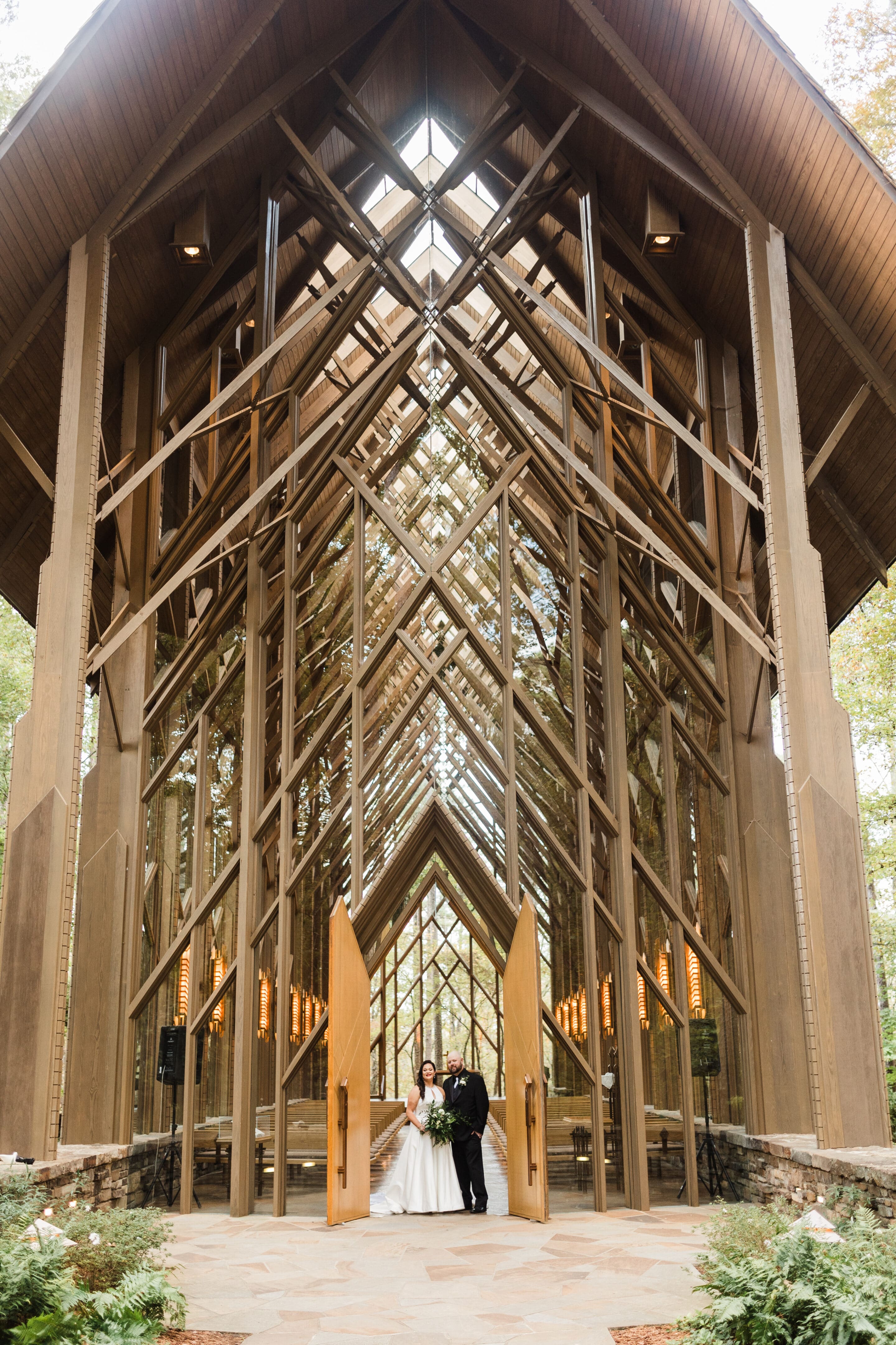 A bride and groom stand at the open doors of a modern glass chapel with wooden beams, surrounded by trees and natural light streaming in through the tall windows.