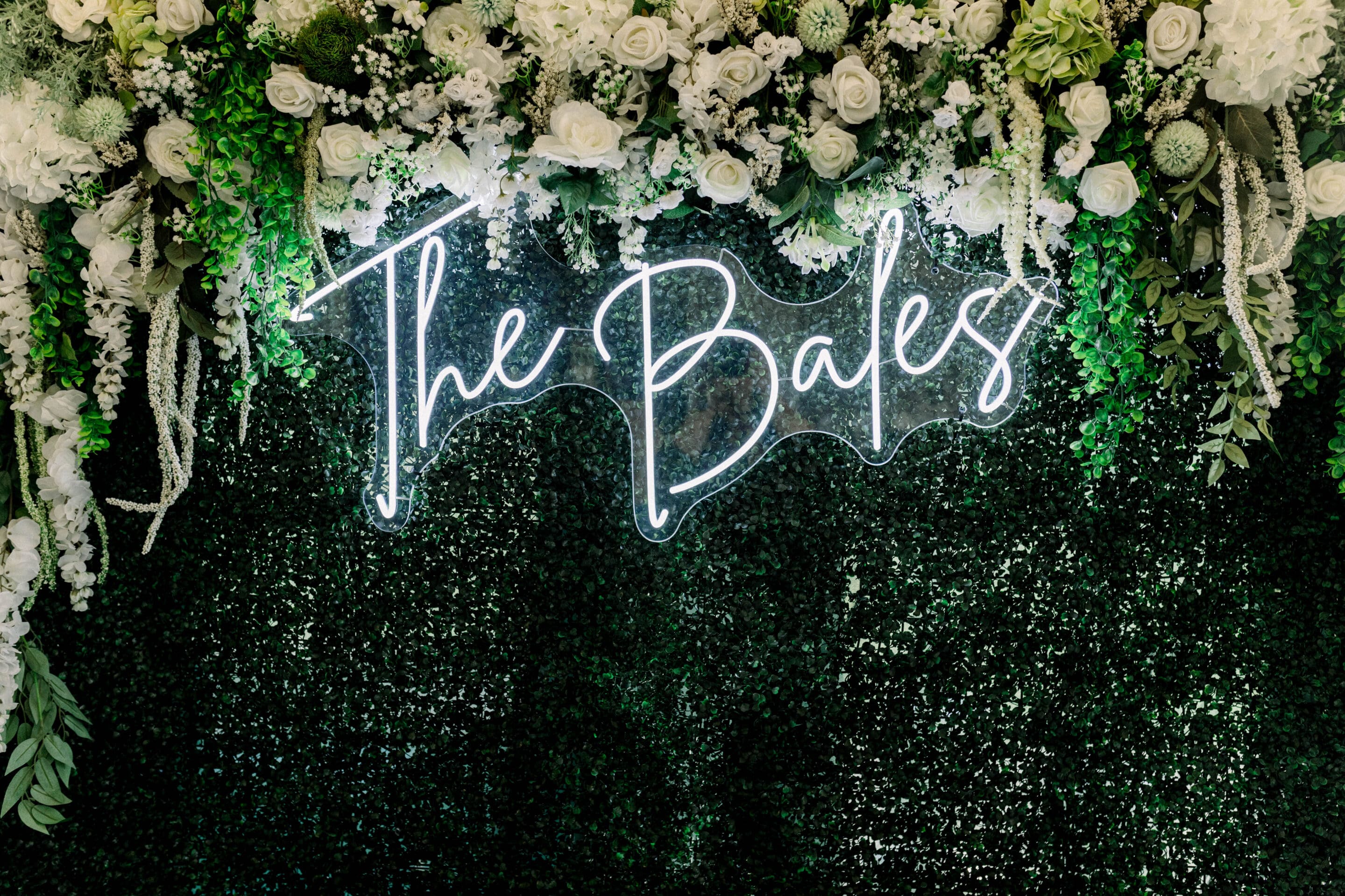 A neon sign reading The Bales is displayed on a lush green faux hedge wall, adorned with white roses, greenery, and floral decorations at the top.