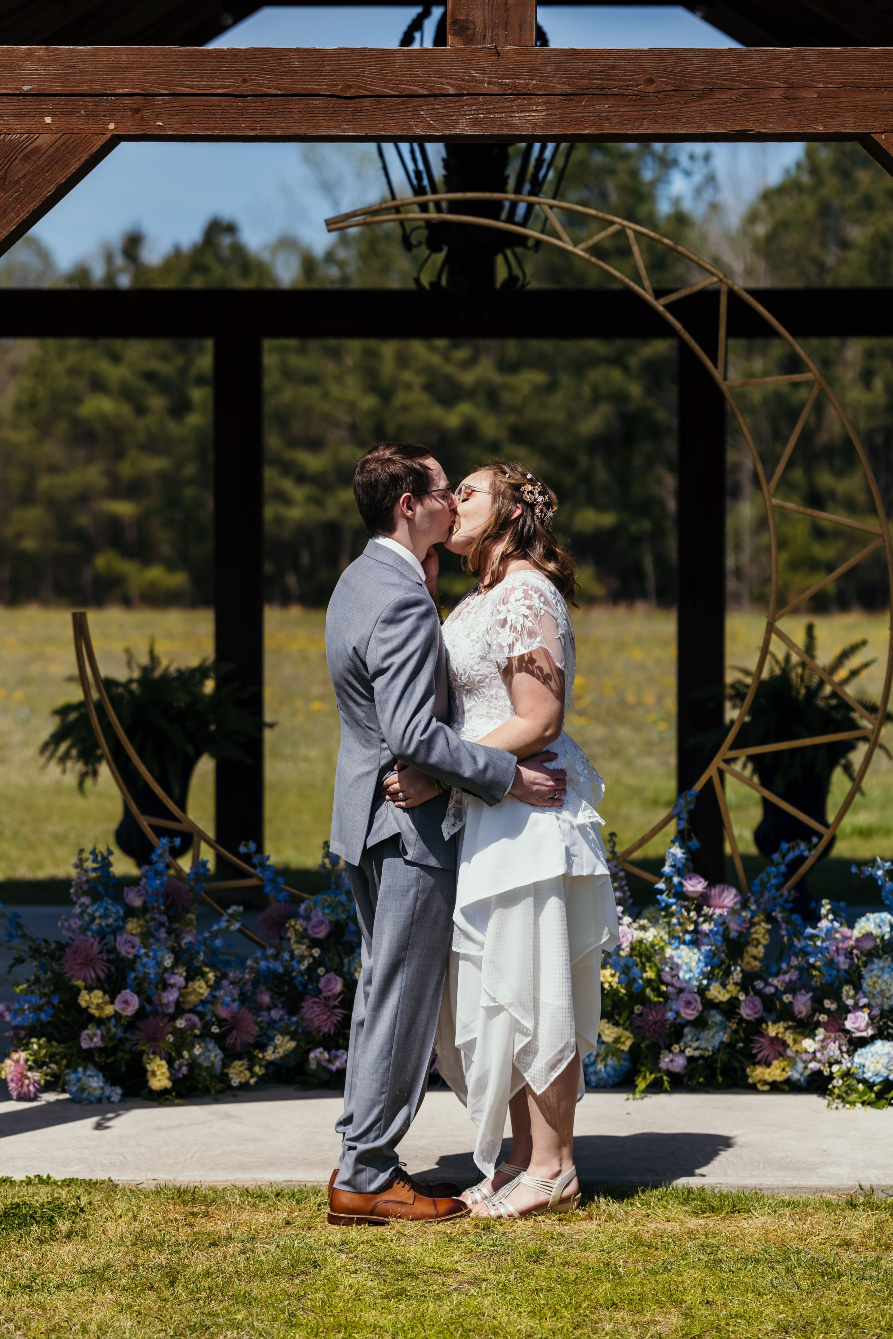 A bride and groom kiss under a wooden arch decorated with flowers and a gold circular frame, surrounded by colorful floral arrangements outdoors on a sunny day.