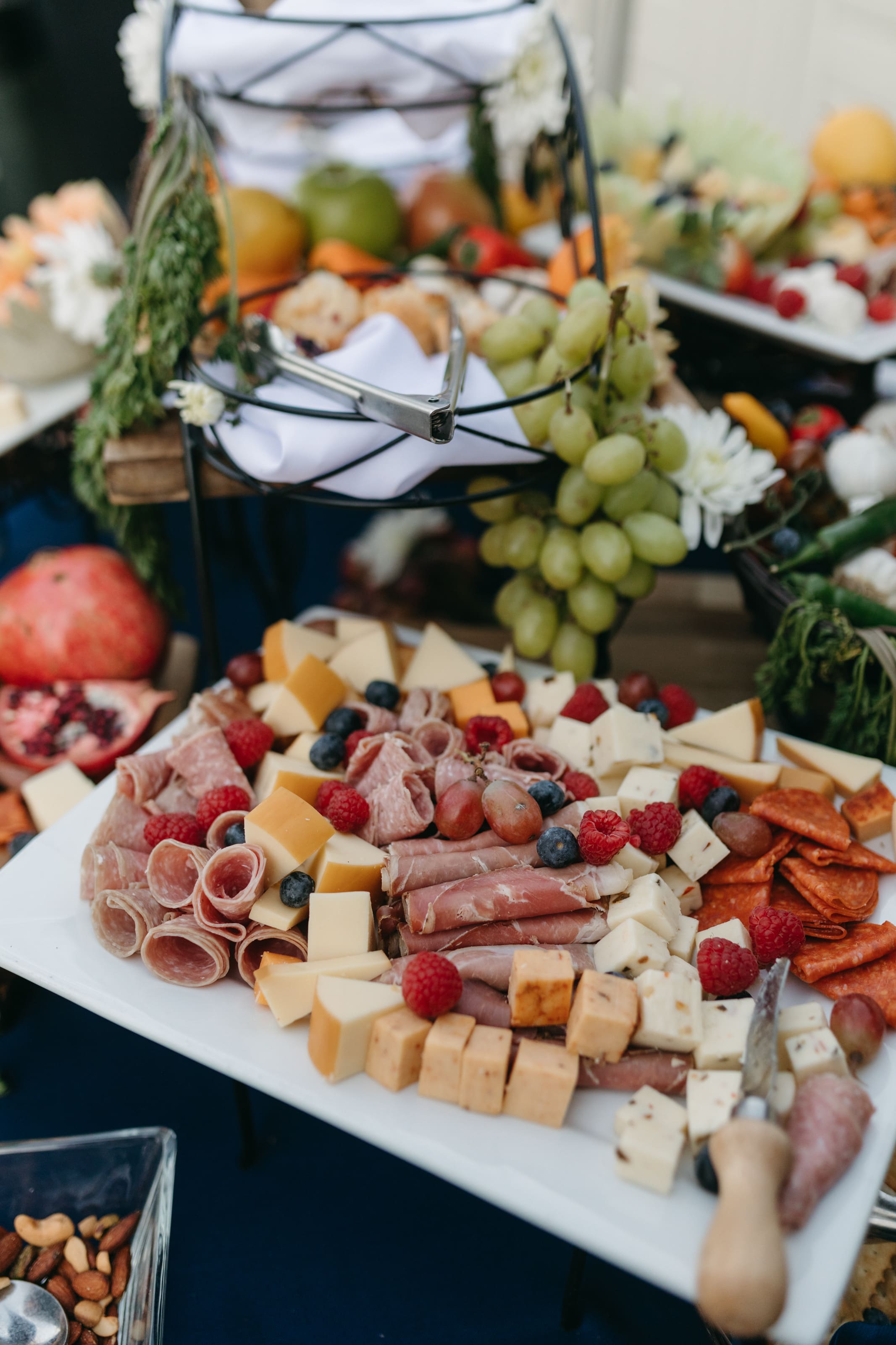 A platter with assorted cheeses, cured meats, berries, and grapes sits on a table, surrounded by pomegranate halves, nuts, and more fruit, creating an inviting charcuterie spread.