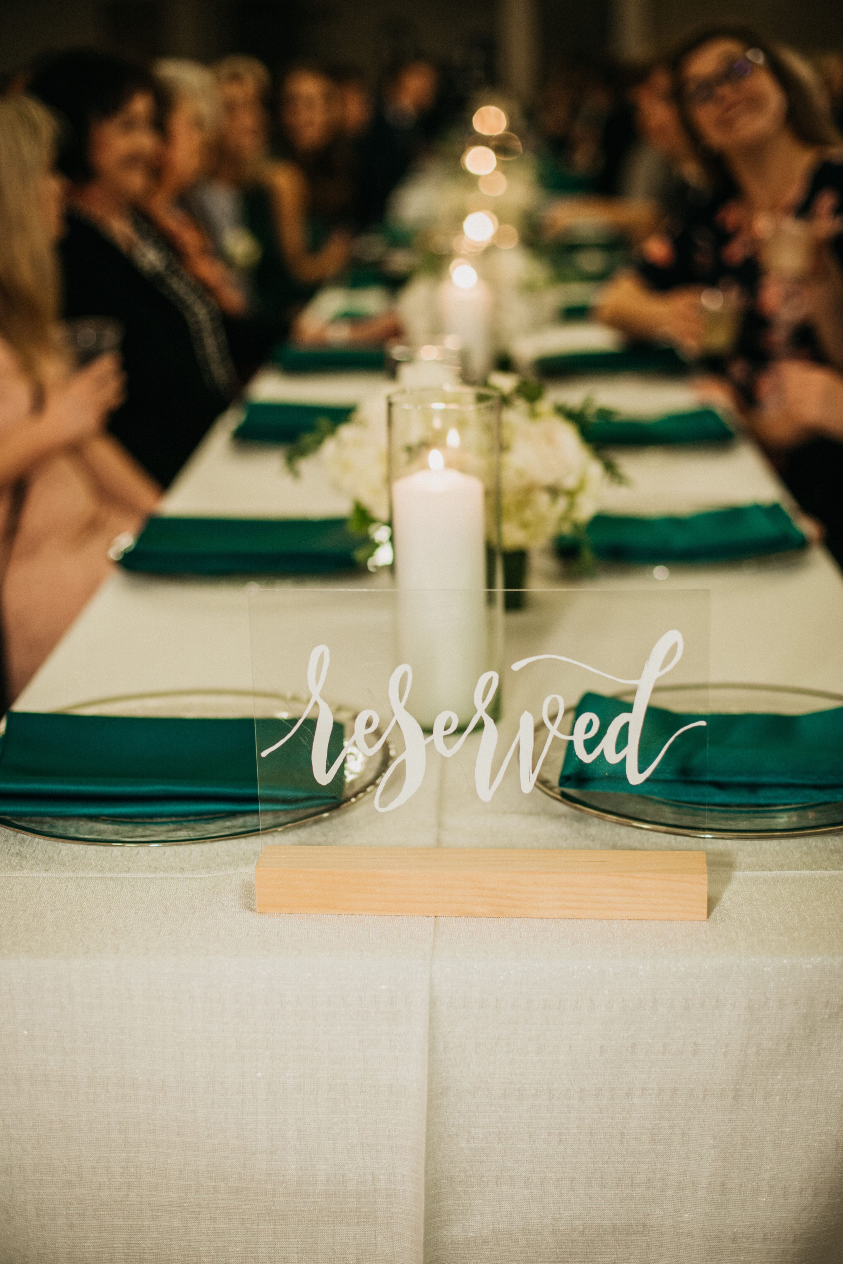 A long table set for an event with teal napkins, candles, and white flowers. A clear acrylic “reserved” sign is displayed at the table’s center. People are seated along both sides, blurred in the background.