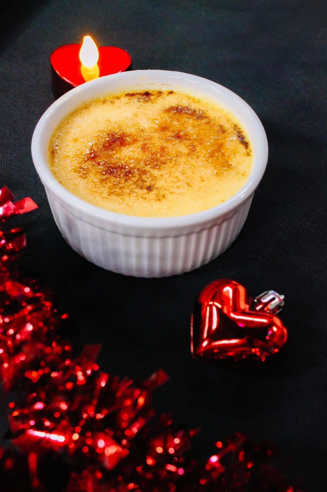 A ramekin of crème brûlée on a dark surface, with a lit red candle, a shiny red heart ornament, and red tinsel decorations nearby.
