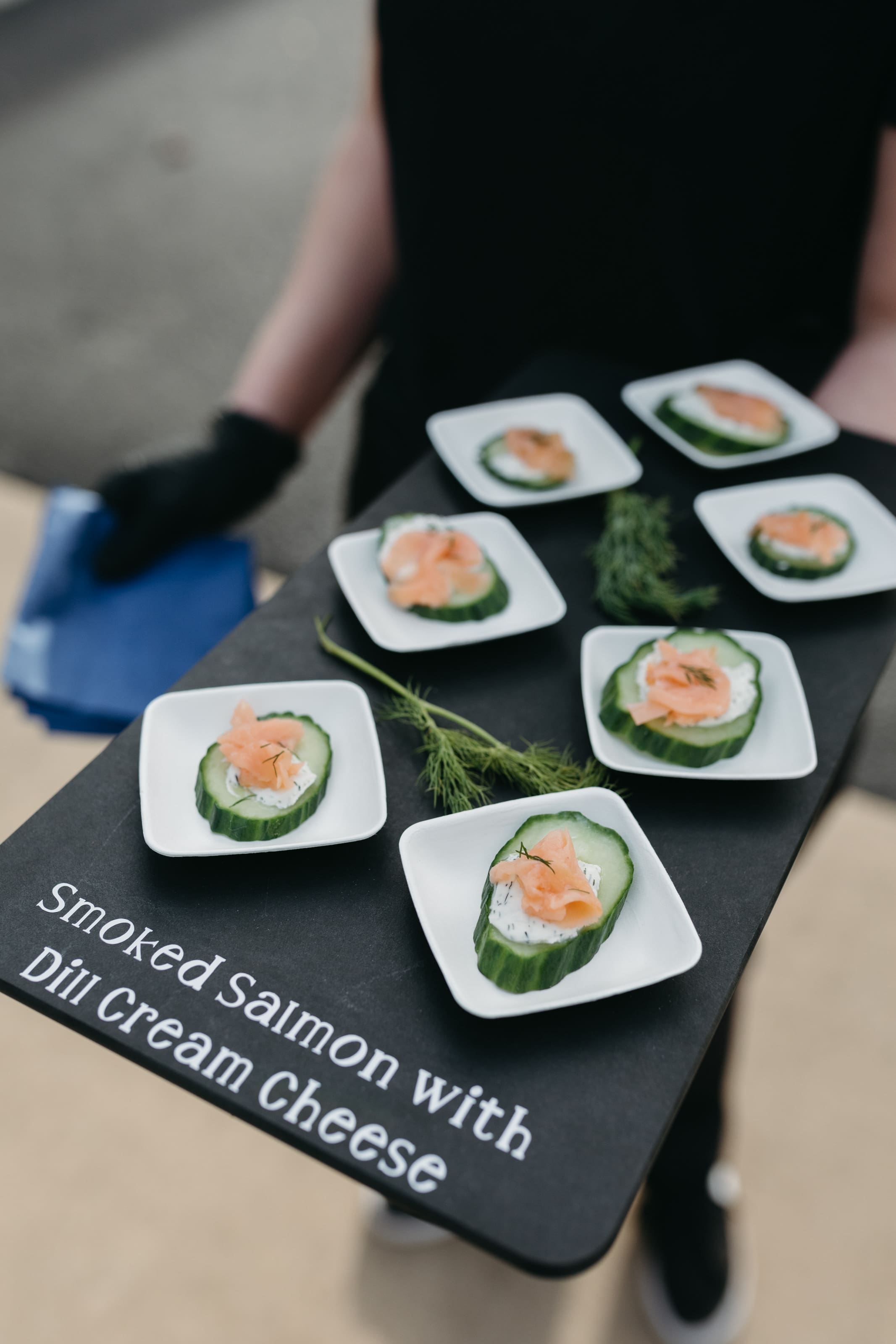 A server holds a tray with small plates of smoked salmon and dill cream cheese on cucumber slices, garnished with fresh dill. The tray text reads, Smoked Salmon with Dill Cream Cheese.