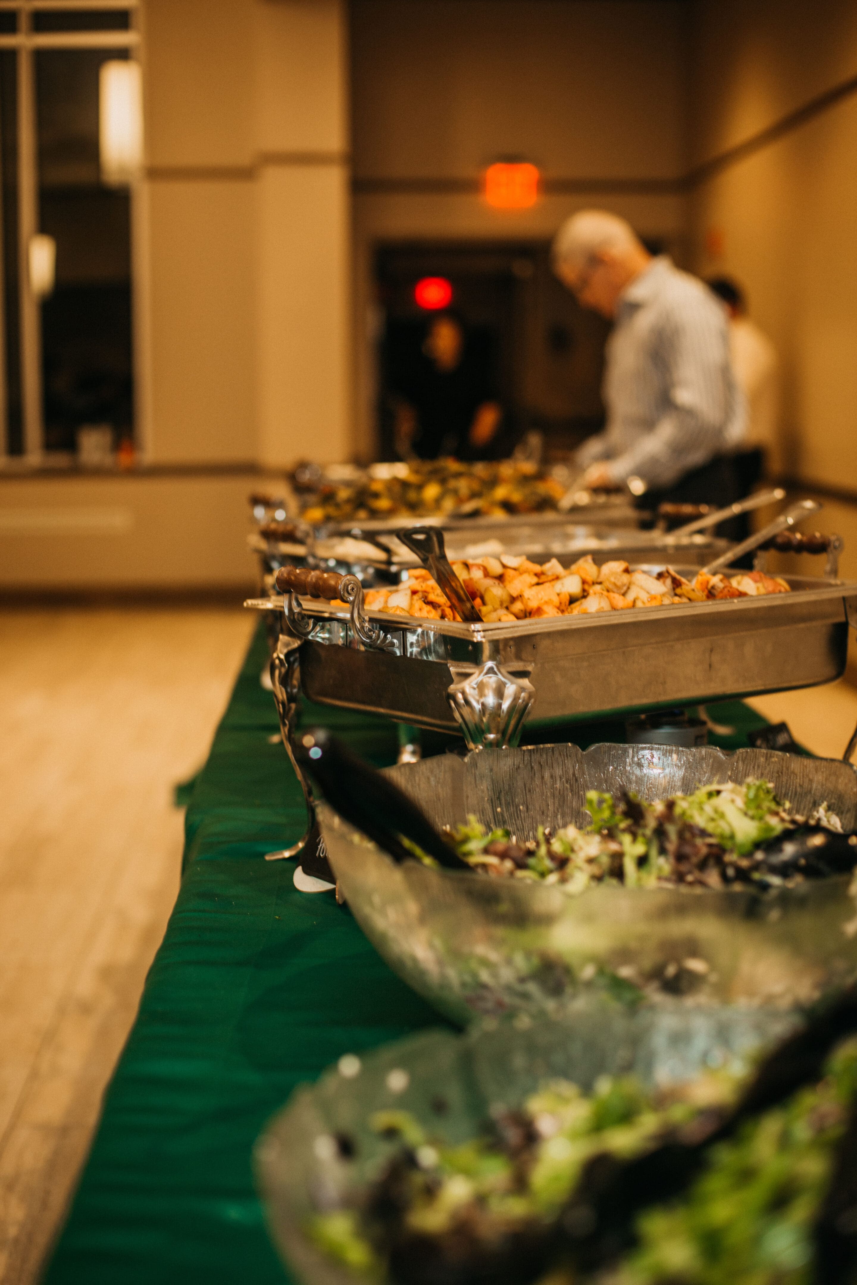 A buffet table with trays of hot food and bowls of salad, set up in a warmly lit room. A person in the background is serving themselves from the buffet.