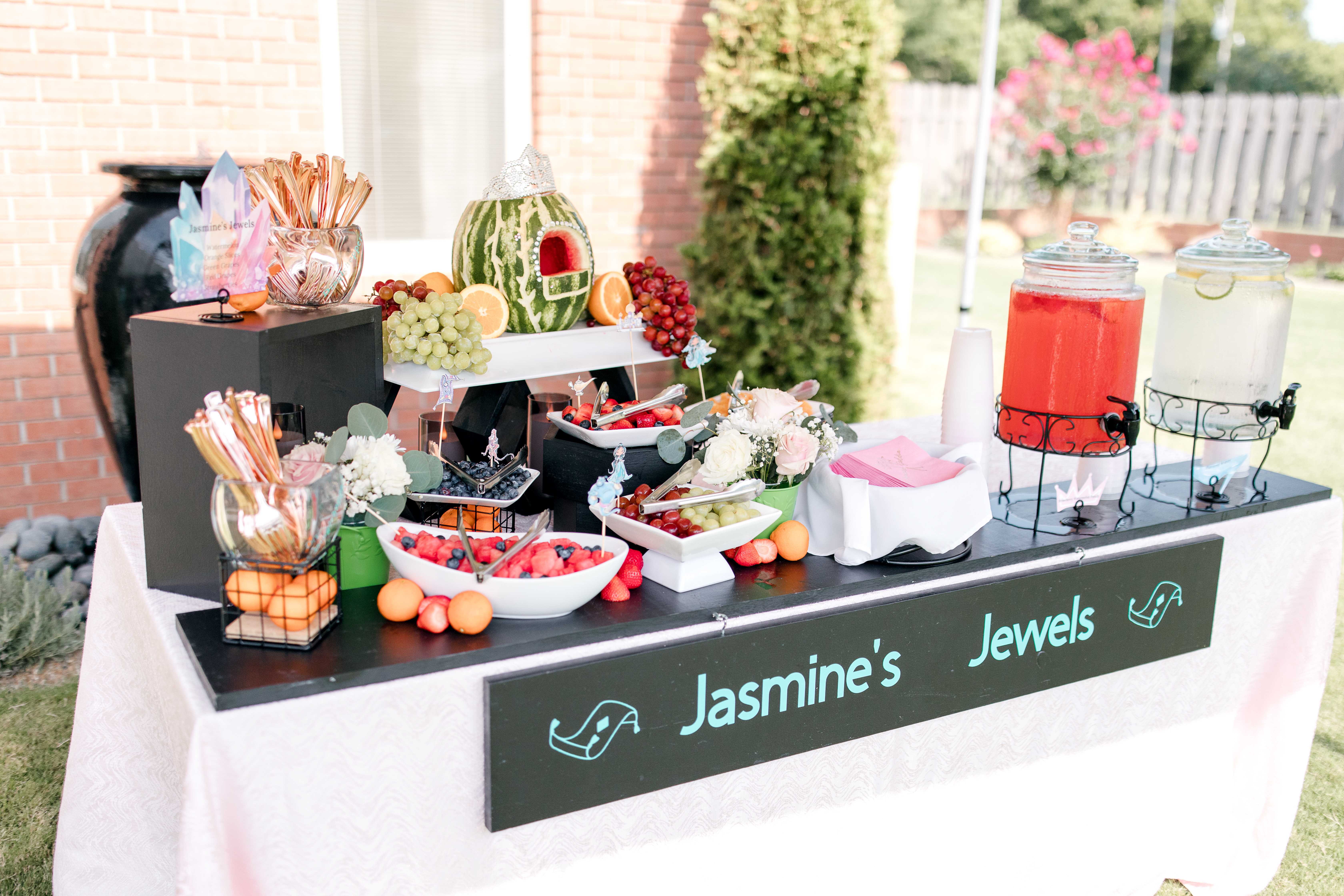 A decorated table labeled Jasmines Jewels displays colorful fruits, a watermelon carved like a house, drinks in dispensers, flowers, and party snacks—perfect Poolside Princess Party Food set outdoors with lush greenery in the background.