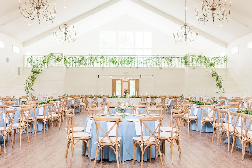 A bright, elegant banquet hall with high ceilings, wooden chandeliers, and large windows. Round tables covered with light blue tablecloths and wooden chairs are arranged neatly. Greenery decorates railings and centerpieces.