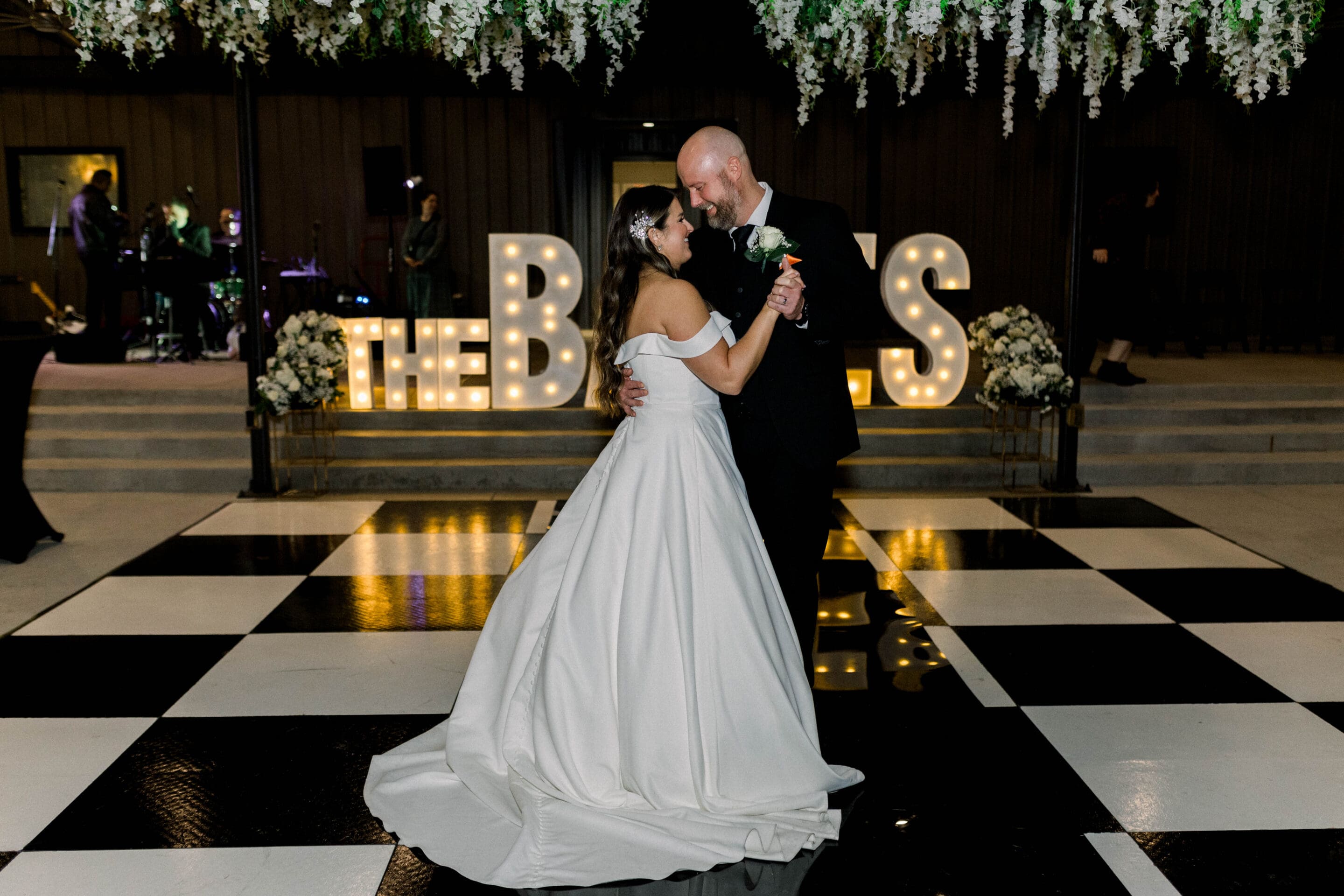 A bride and groom share a first dance on a black-and-white checkered floor, with floral arrangements and illuminated THE BOSS letters in the background. White flowers hang overhead, and a band performs off to the side.