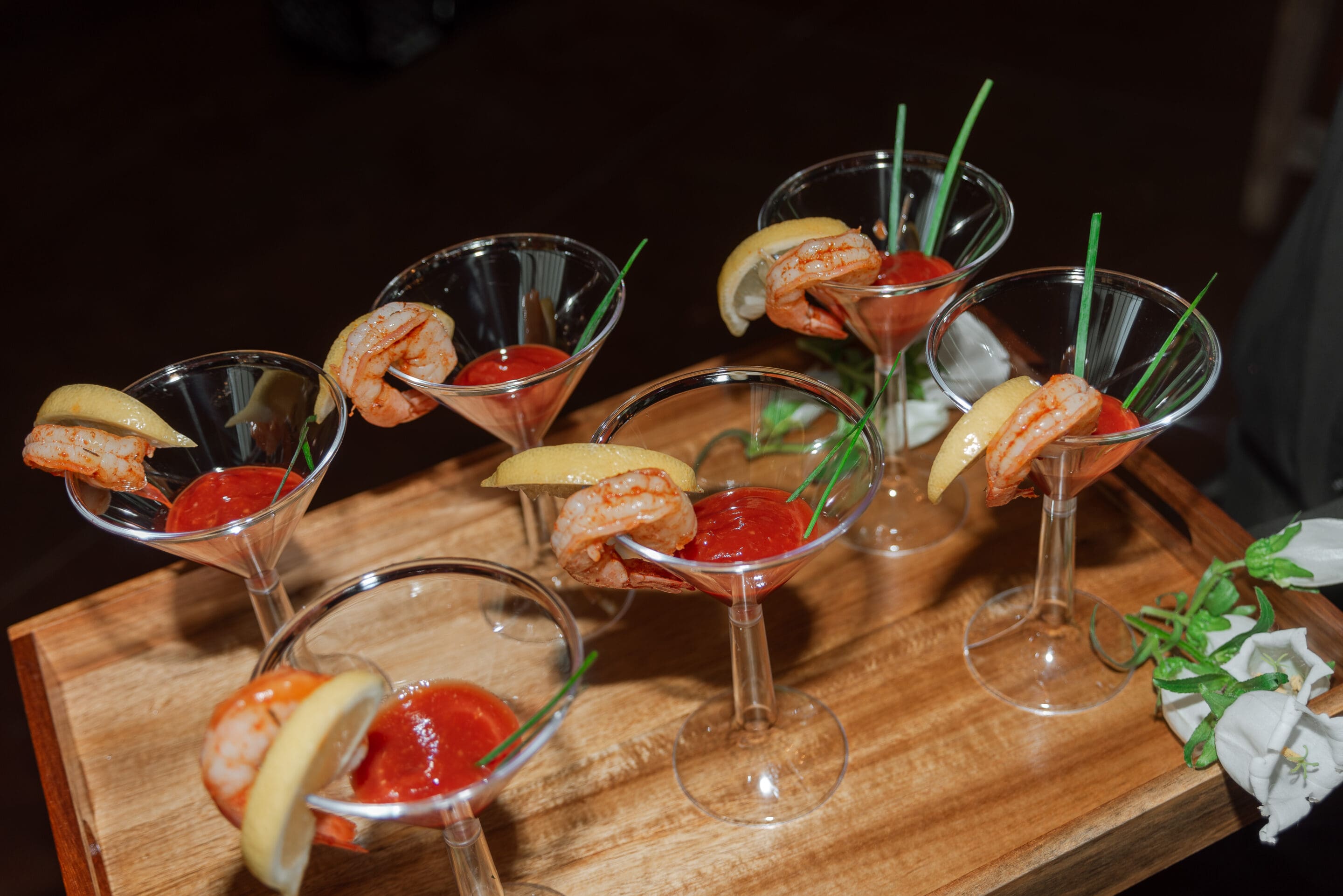 Six martini glasses filled with cocktail sauce, each garnished with a shrimp, a lemon wedge, and a piece of chive, are arranged on a wooden tray. White flowers are visible at the corner of the tray.