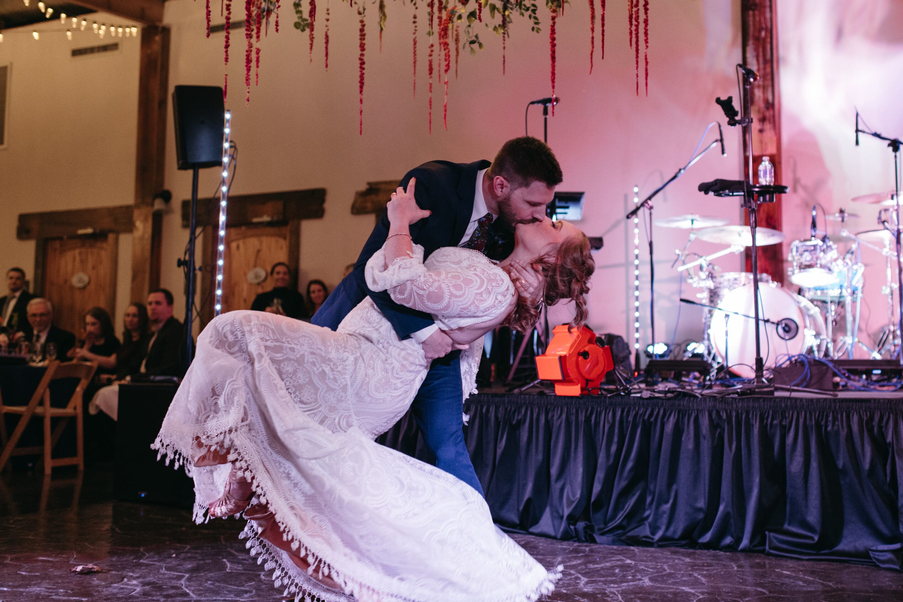 A bride and groom share a dramatic dip and kiss on the dance floor during their wedding reception, with guests watching and a stage with musical instruments in the background.