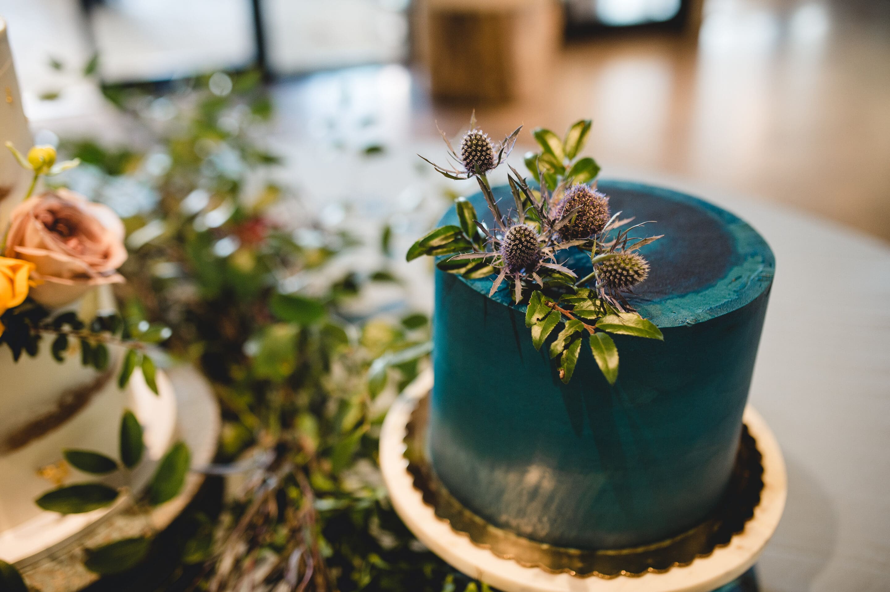 A round teal cake with a smooth finish is decorated with small purple thistles and green leaves, sitting on a gold-edged cake board. Greenery is scattered on the surface near the cake.
