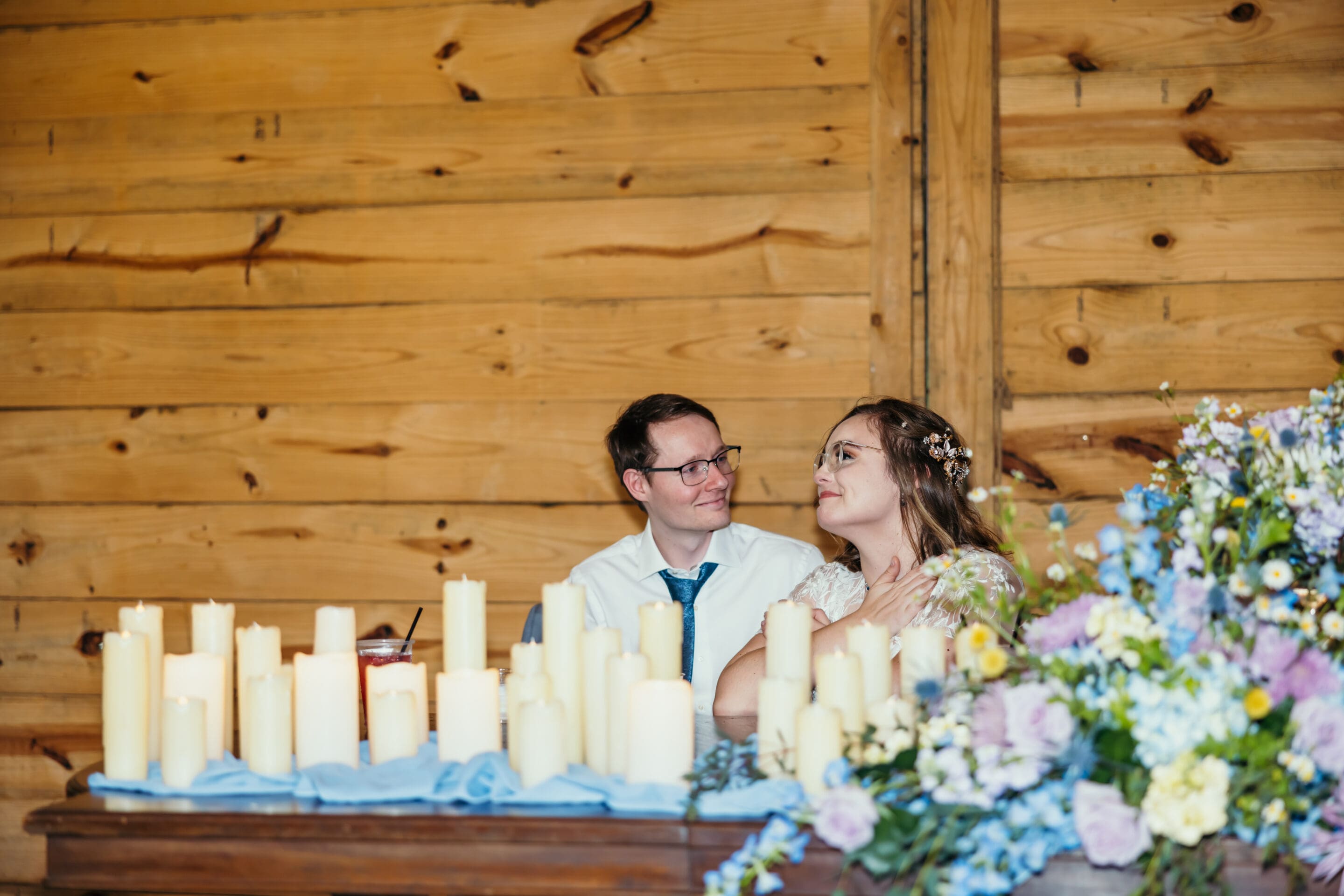 A bride and groom sit together at a table decorated with candles and pastel flowers, smiling at each other, in front of a wooden wall.