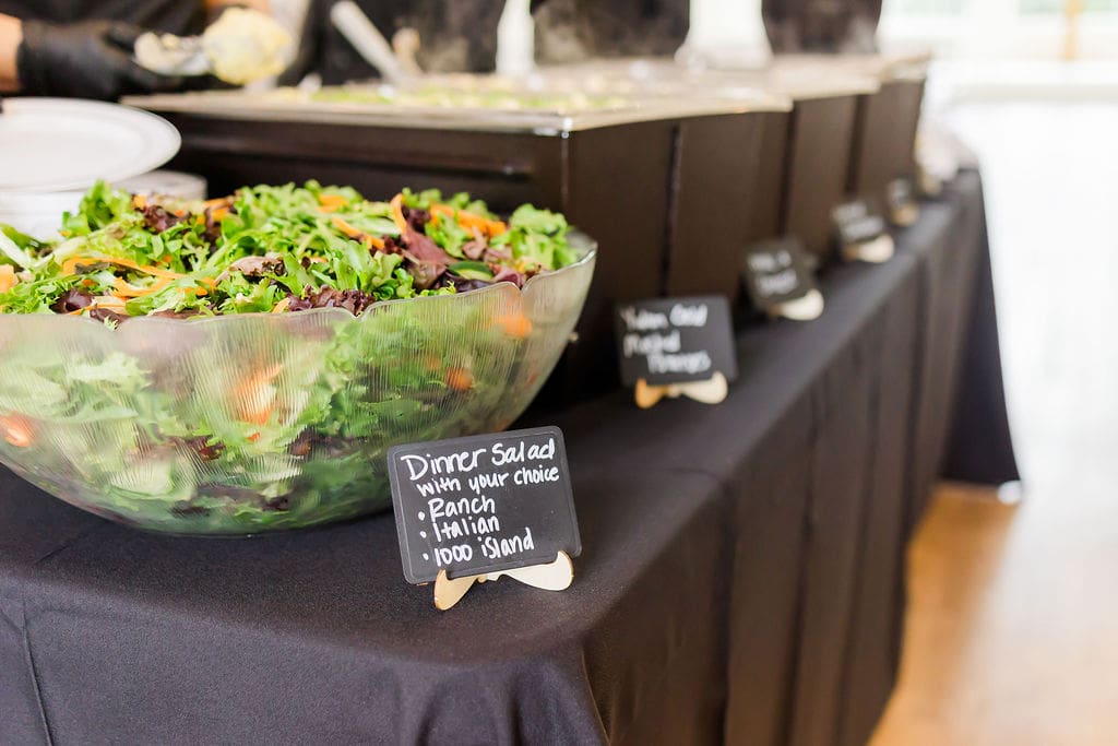 A large bowl of mixed green salad sits on a black tablecloth next to a buffet with menu cards, including one for Dinner Salad offering Ranch, Italian, and Thousand Island dressings.