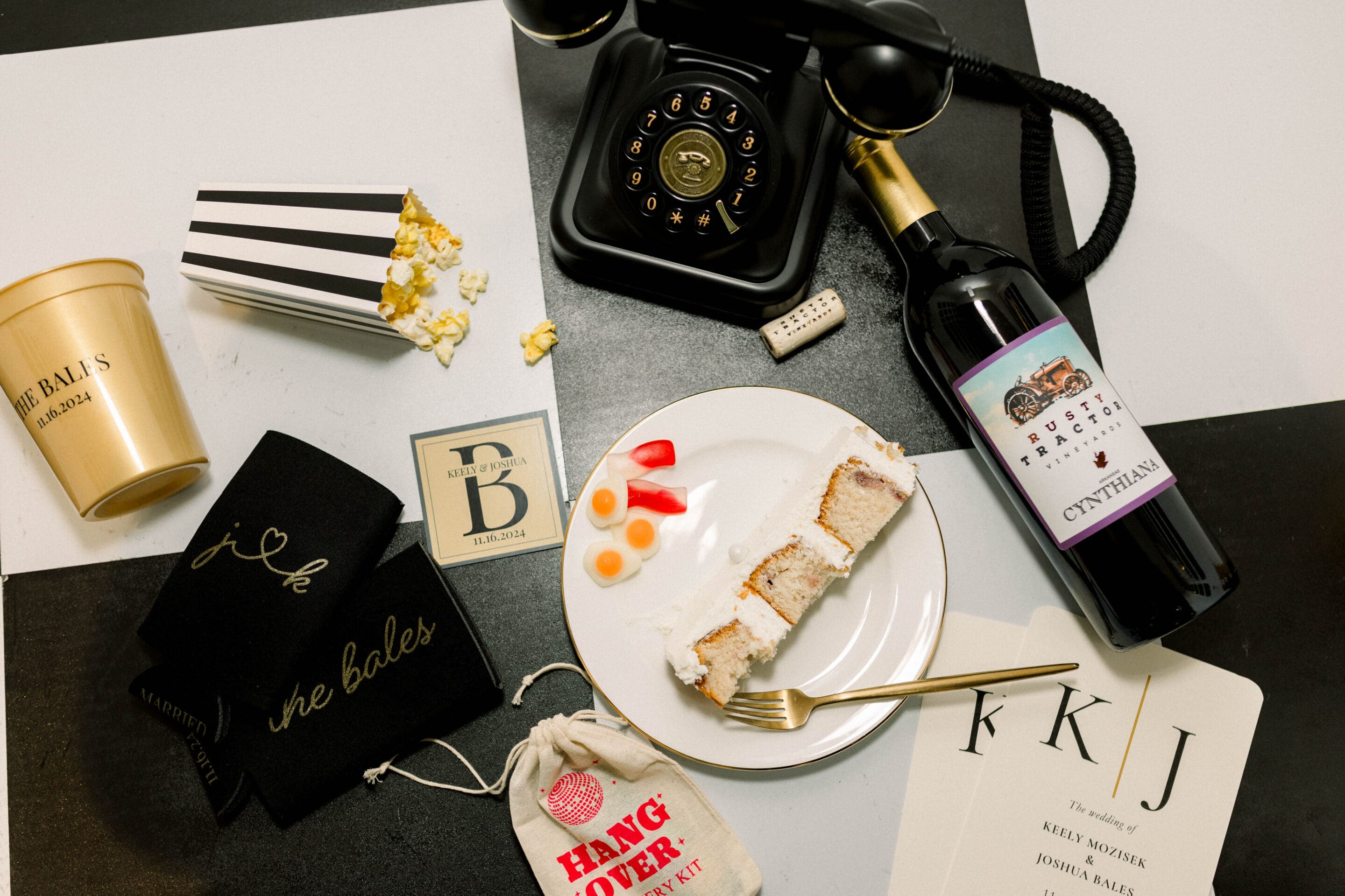 A table with cake, wine, popcorn, a gold cup, a black rotary phone, a domino, a black napkin, wedding stationery, and a small hangover kit pouch on a black-and-white checkered surface.