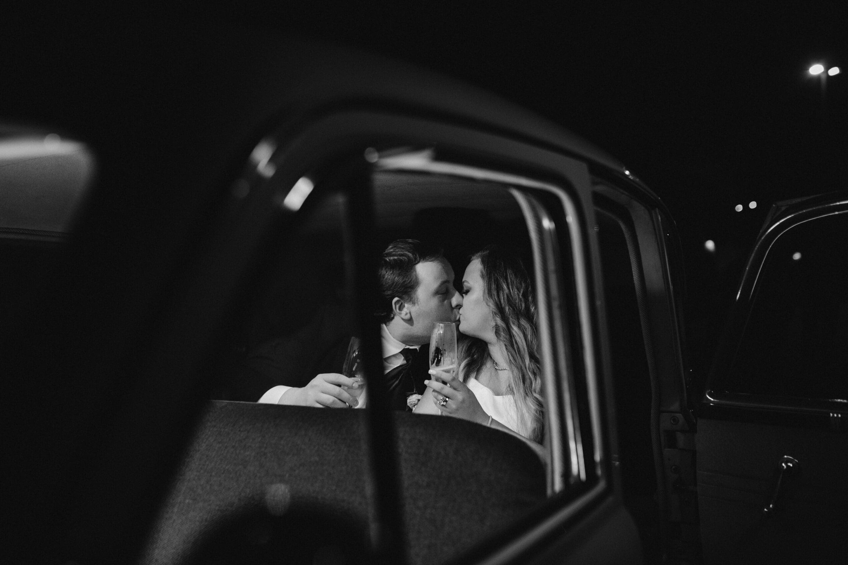 A black-and-white photo shows a couple dressed in formal wear, sharing a kiss and holding champagne glasses while sitting in the backseat of a car at night. The image is taken from outside the open car door.