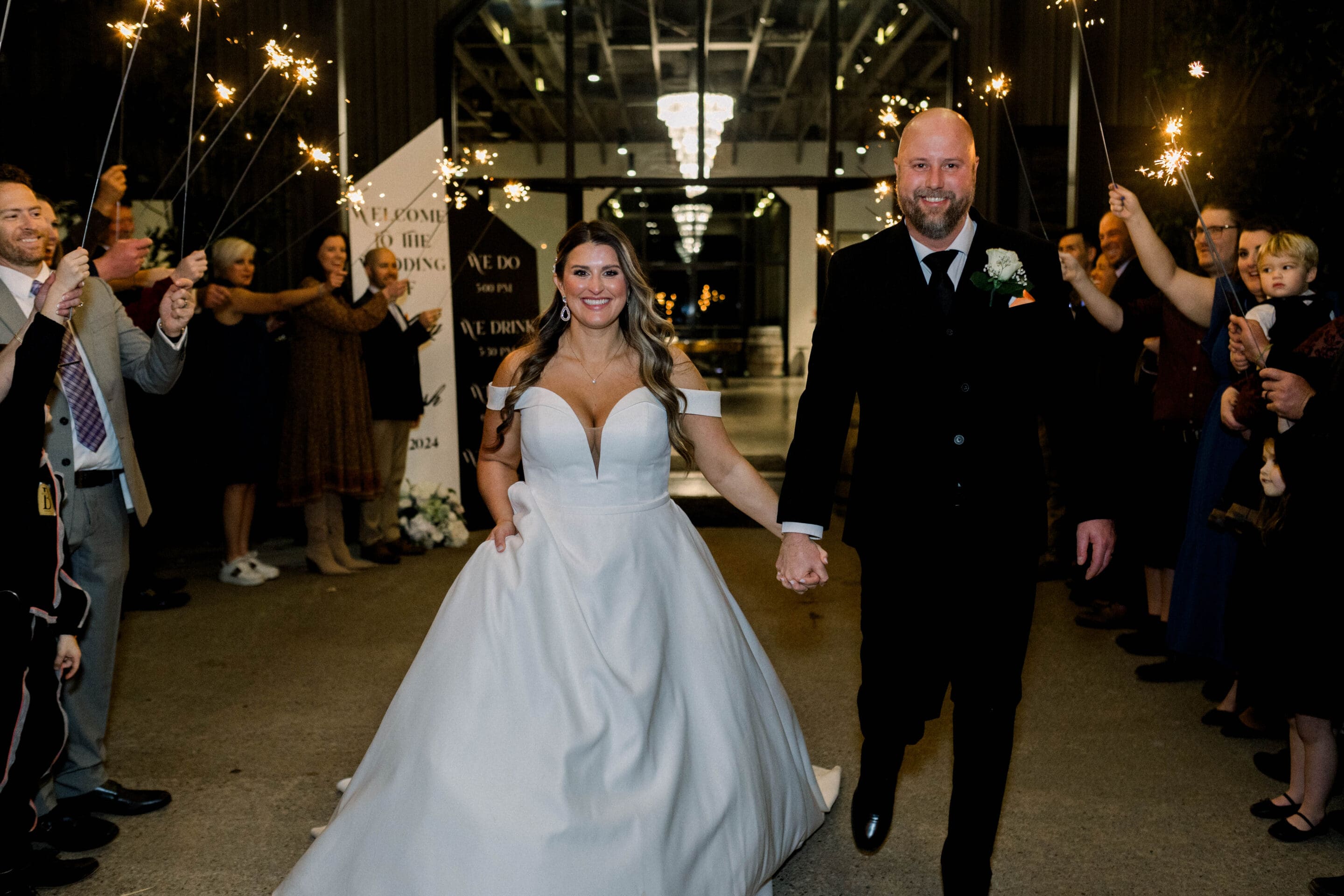 A bride in a white gown and a groom in a black suit walk hand-in-hand, smiling, through a crowd of guests holding sparklers at night in a decorated indoor venue.