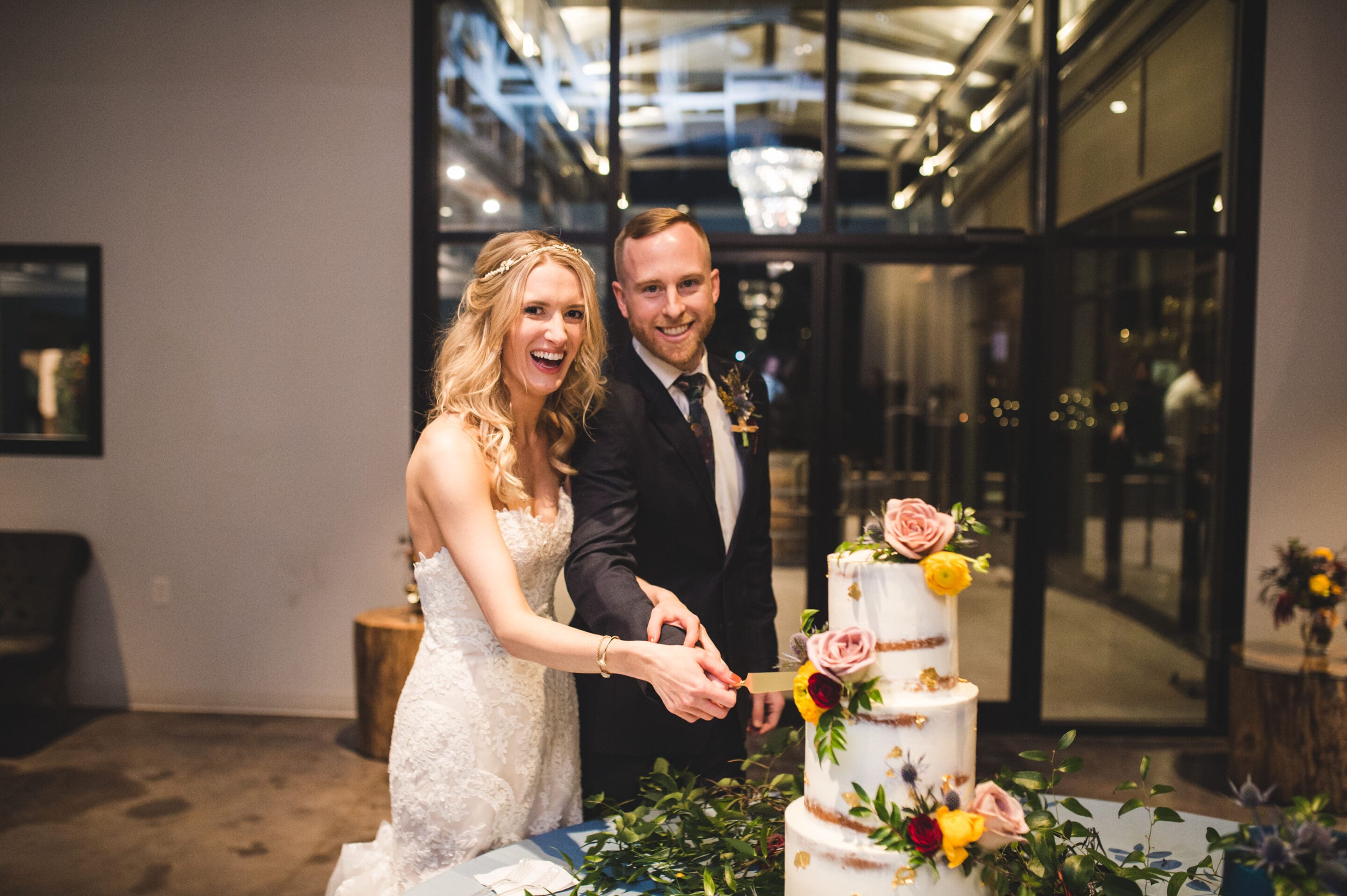 A bride and groom smile joyfully as they cut their wedding cake together. The three-tiered cake is decorated with fresh flowers and greenery. They stand in a modern, warmly lit venue with large glass doors behind them.