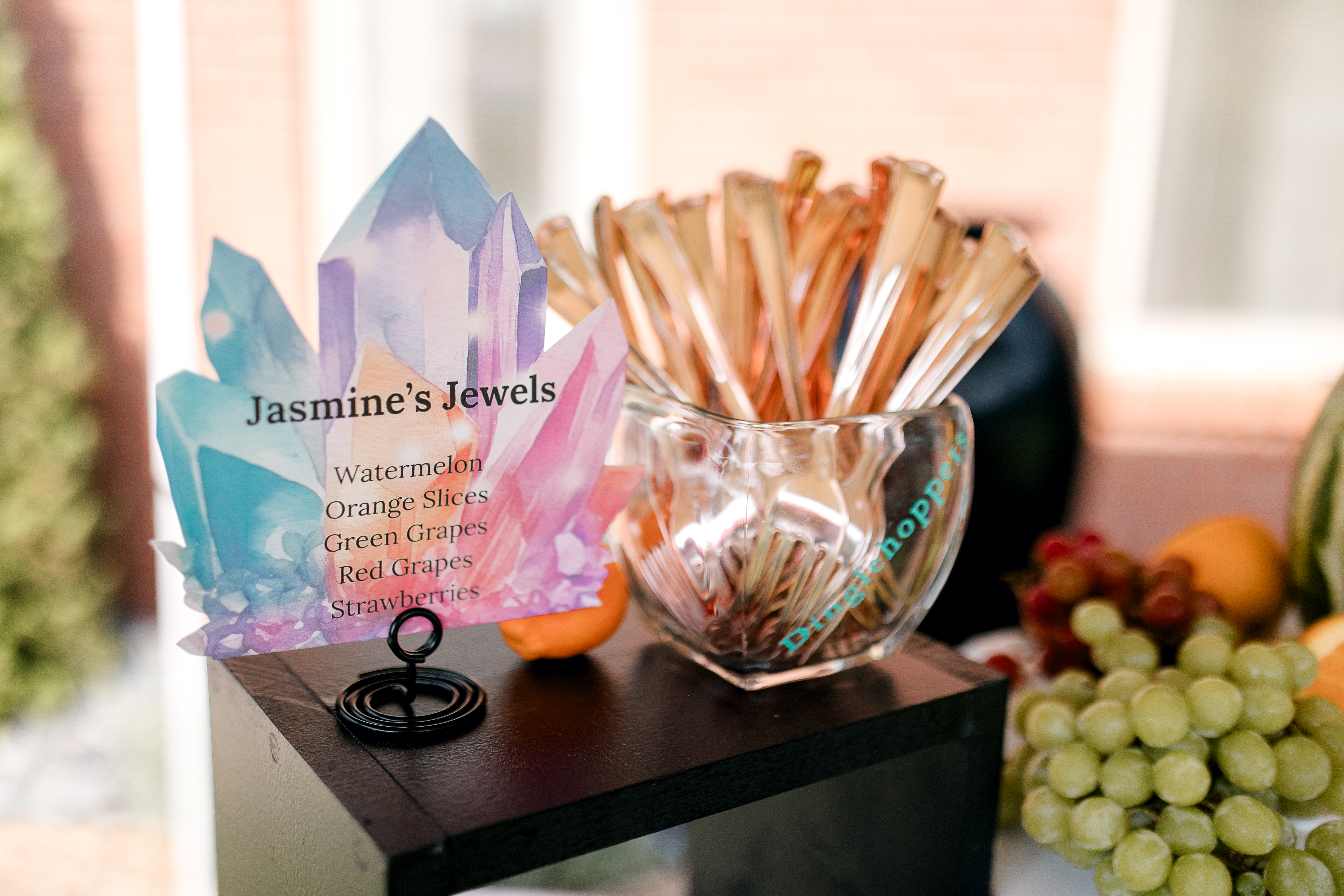 A colorful sign labeled Jasmines Jewels lists fruit options next to a glass cup holding rose-gold forks, perfect for Poolside Princess Party Food. Green and red grapes are displayed on the right, with a blurred brick building in the background.
