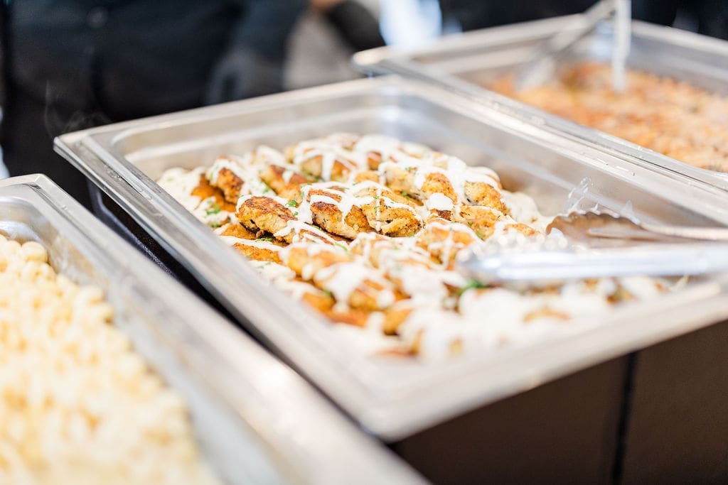 A close-up of a buffet tray filled with breaded chicken pieces drizzled with white sauce, garnished with herbs. Metal tongs rest on the edge of the tray, and other trays of food are visible nearby.