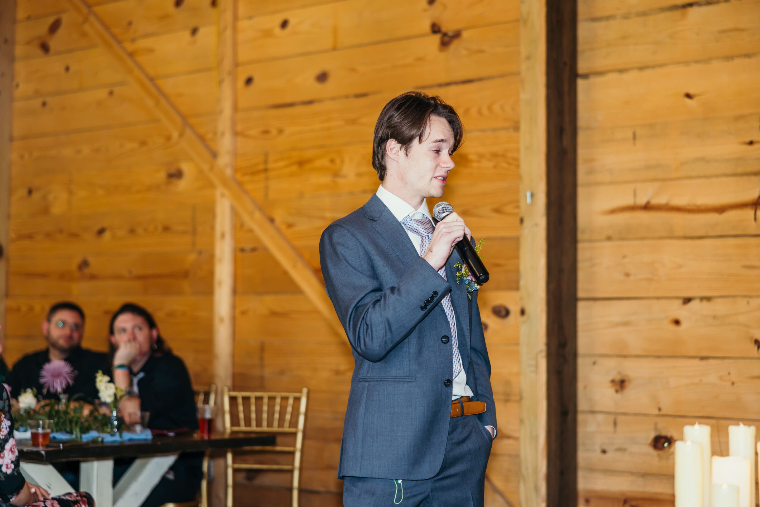 A young man in a gray suit holds a microphone and speaks in a wooden-paneled room, while people seated at a table in the background listen attentively.