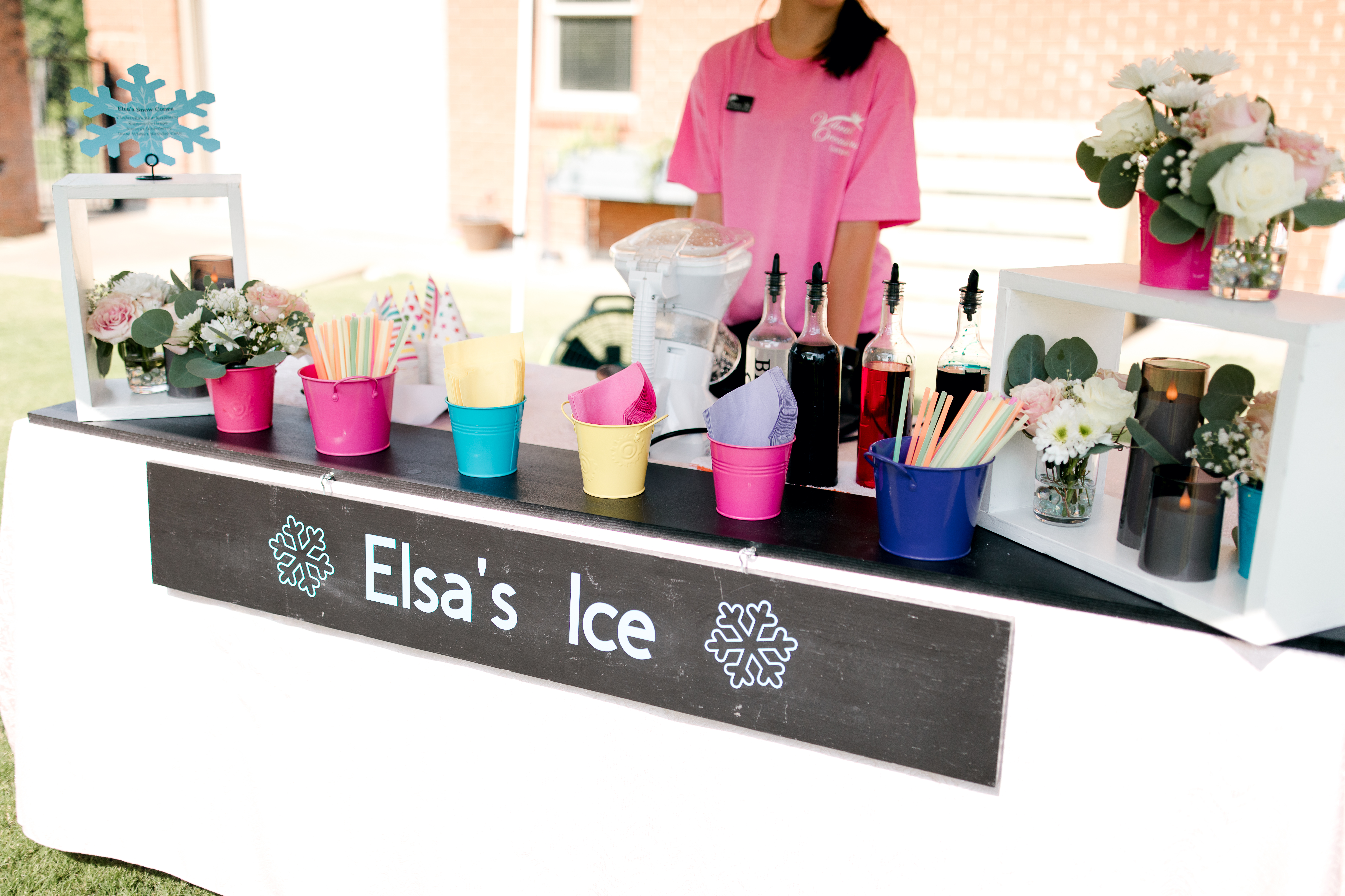 A colorful ice stand labeled Elsas Ice displays syrups, cups, straws, and flowers on a white table with a staff member in a pink shirt—perfect Poolside Princess Party Food for your next celebration.