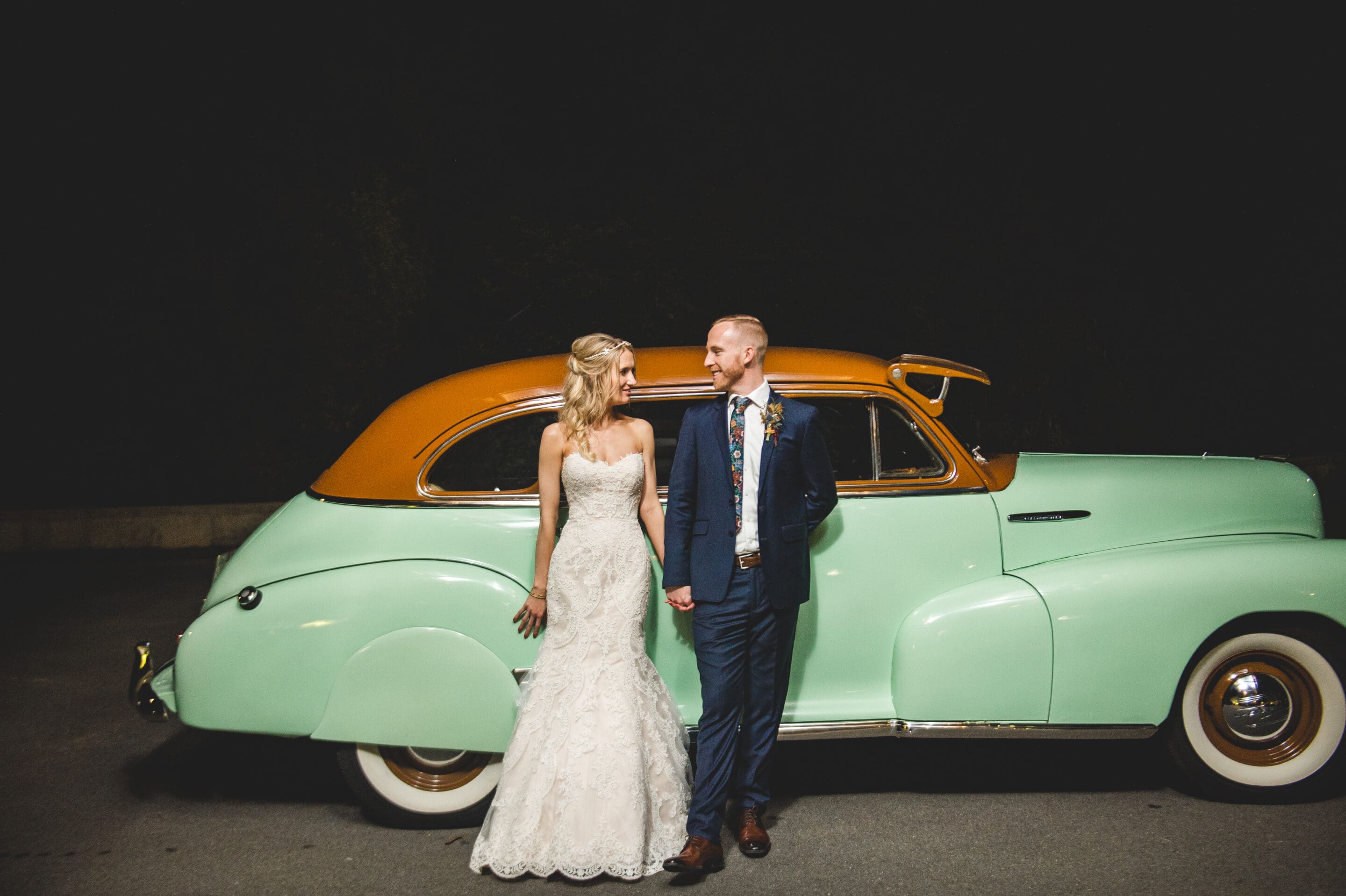 A bride in a white gown and a groom in a blue suit stand holding hands in front of a vintage mint green and tan car at night, both smiling at each other.