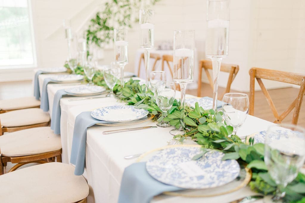 A long, elegant dining table set with blue and white patterned plates, glasses, silverware, light blue napkins, and a lush green garland centerpiece with tall glass candle holders. Wooden chairs surround the table.