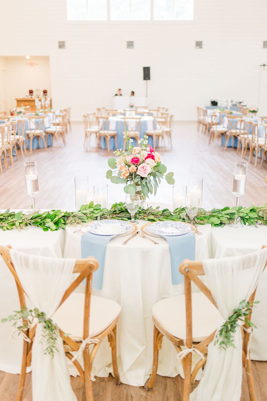 A bright, airy wedding reception hall with round tables, wooden chairs draped in white fabric, greenery and candles, and a central sweetheart table decorated with a pink and white floral arrangement.