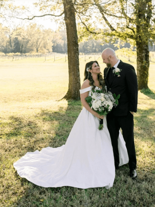 Arkansas Wedding Couple A bride in a white dress holds a bouquet and smiles at a groom in a black suit. They stand together outdoors on grass, near trees, with sunlight streaming through the branches.