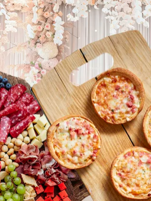 147 A charcuterie board with sliced meats, cheese cubes, green grapes, blueberries, and round mini pizzas on a wooden serving board, with a floral background.