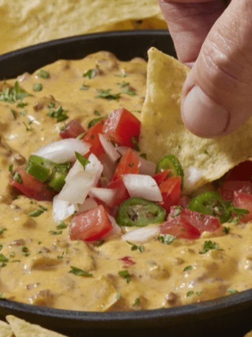 148 A hand dips a tortilla chip into a bowl of cheesy queso dip topped with diced tomatoes, onions, jalapeños, and chopped herbs.