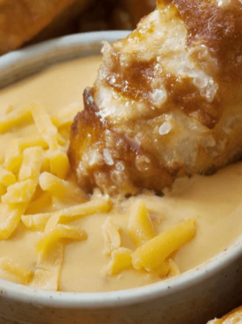 149 A soft pretzel dipped in creamy cheese sauce, garnished with shredded cheddar cheese, served in a small bowl.