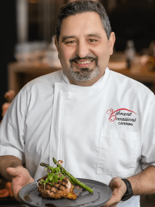 150 A chef in a white jacket with Vibrant Occasions Catering embroidered on it smiles while holding a plate of grilled salmon topped with asparagus and garnish. The background is softly blurred.