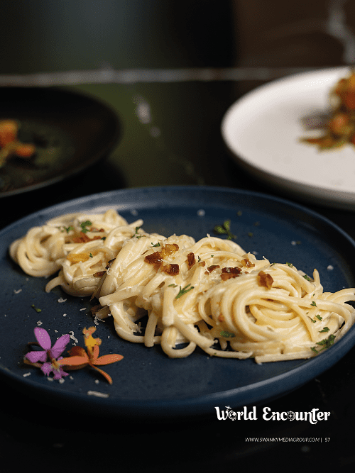 151 A blue plate with creamy pasta garnished with herbs, grated cheese, and small flower blossoms, placed on a dark table with blurred dishes in the background. The words World Encounter are in the bottom right corner.