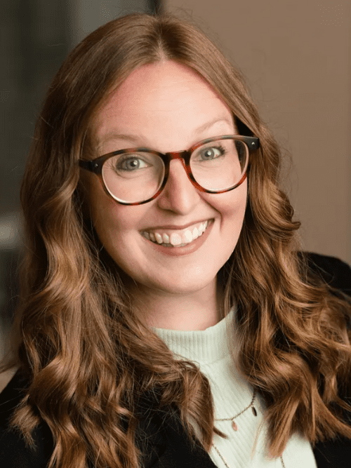 Leah Seale of Vibrant Occasions Catering A woman with long, wavy light brown hair and glasses smiles at the camera. She is wearing a light green top and a black jacket, with a blurred indoor background.