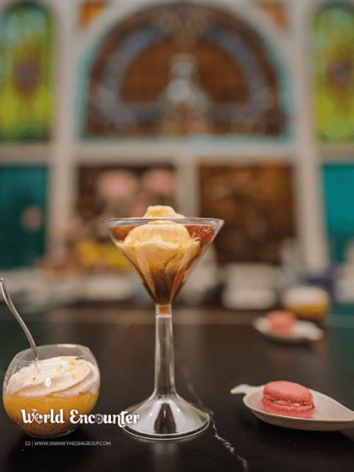 Fancy Desserts Catering Arkansas A glass of affogato sits on a dark counter, accompanied by a dessert in a small cup with a spoon and a pink macaron on a plate. Stained glass windows are blurred in the background. Text reads “World Encounter.”.