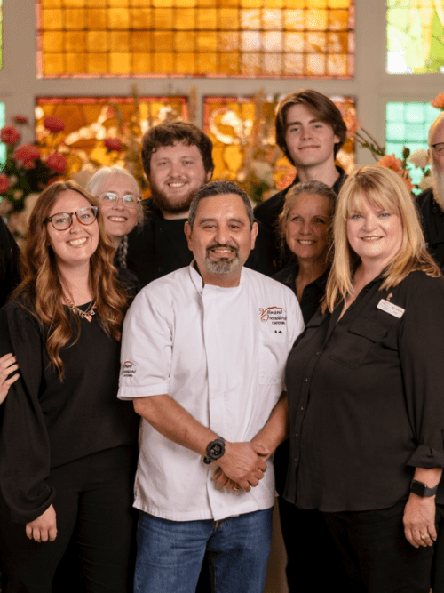 Vibrant Occasions Catering Staff A group of people, including a chef in a white coat, pose together and smile in front of stained glass windows and floral arrangements. Most are wearing black clothing.